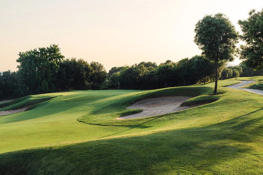 A lush green golf course with sand bunkers, trees, and a path winding through the landscape during sunset or late afternoon.