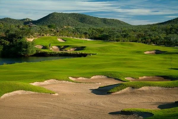 A scenic golf course with green fairways, sand bunkers, water hazards, and a backdrop of lush trees and hills under a partly cloudy sky.