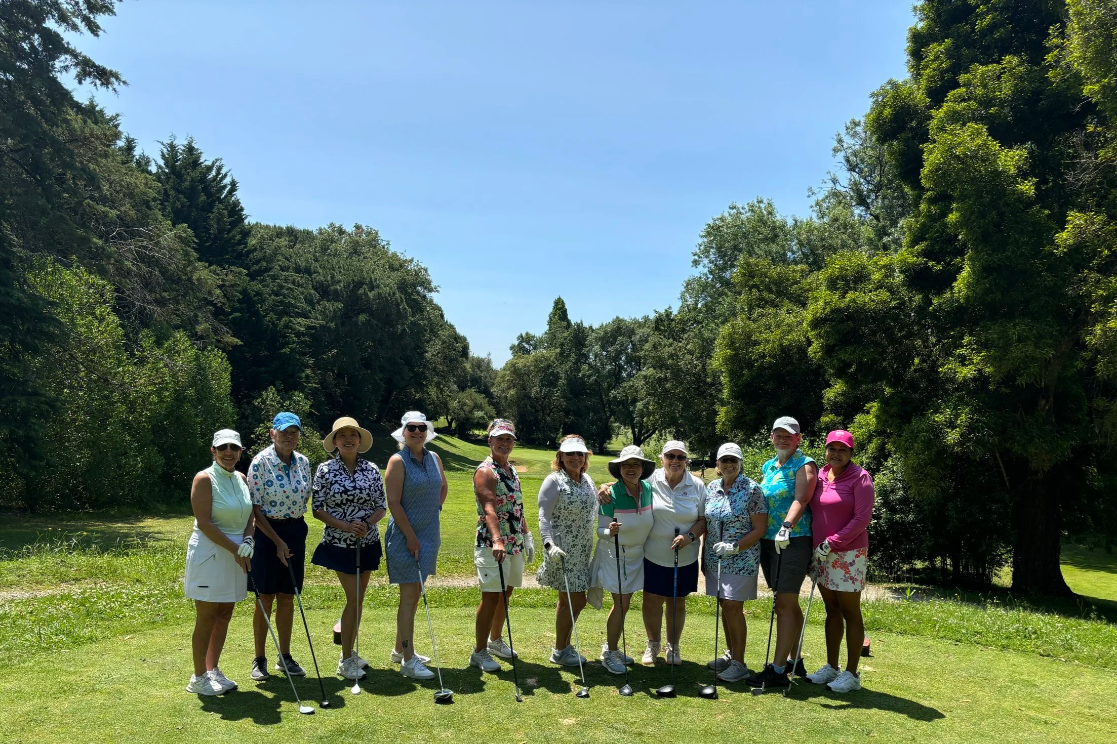 Group of women posing on a golf course in sunny weather, holding golf clubs, wearing hats and sunglasses.