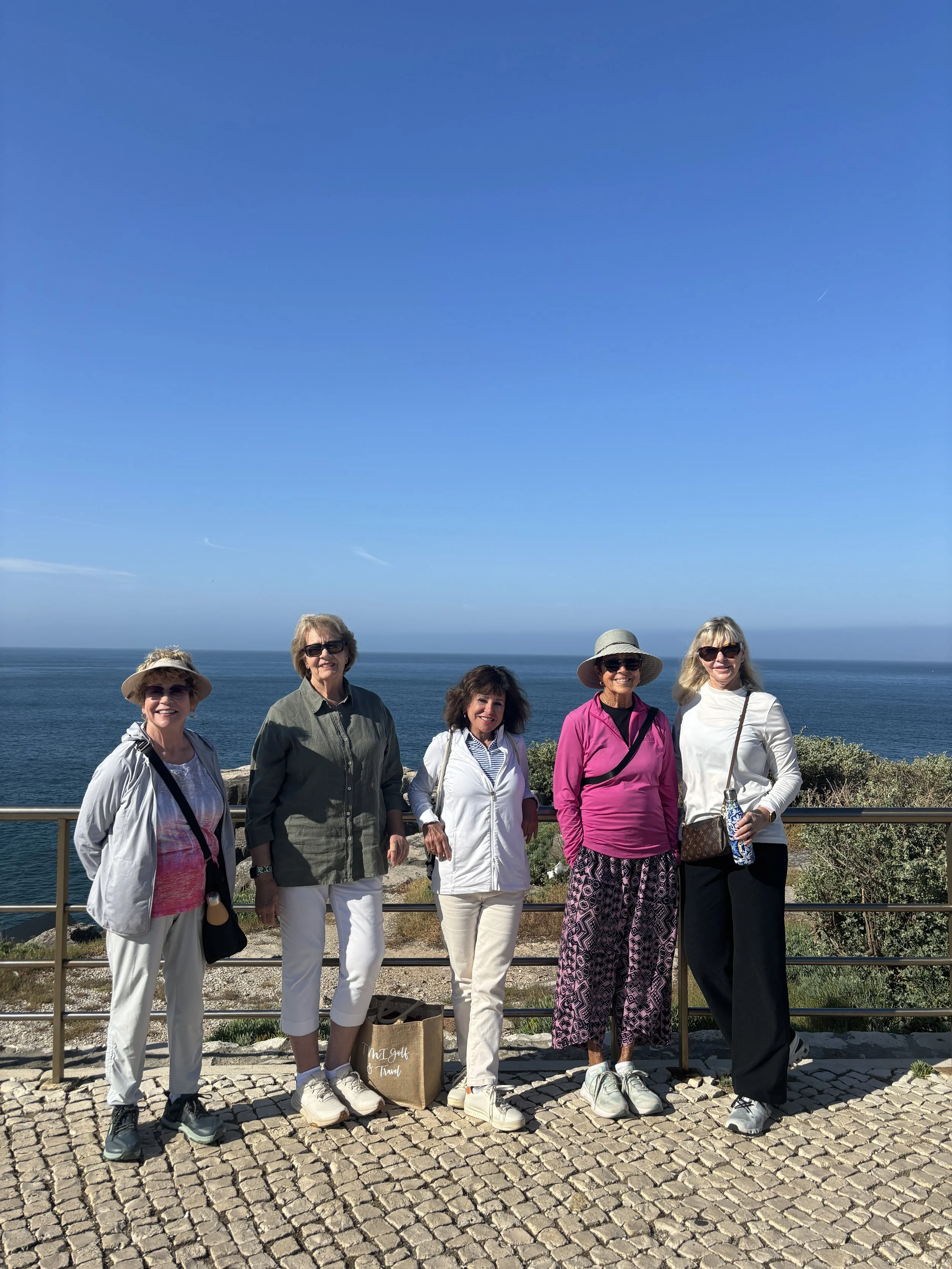 Group of five women standing outdoors on a cobblestone path with a metal railing, posing for a photo with the ocean and clear blue sky in the background.