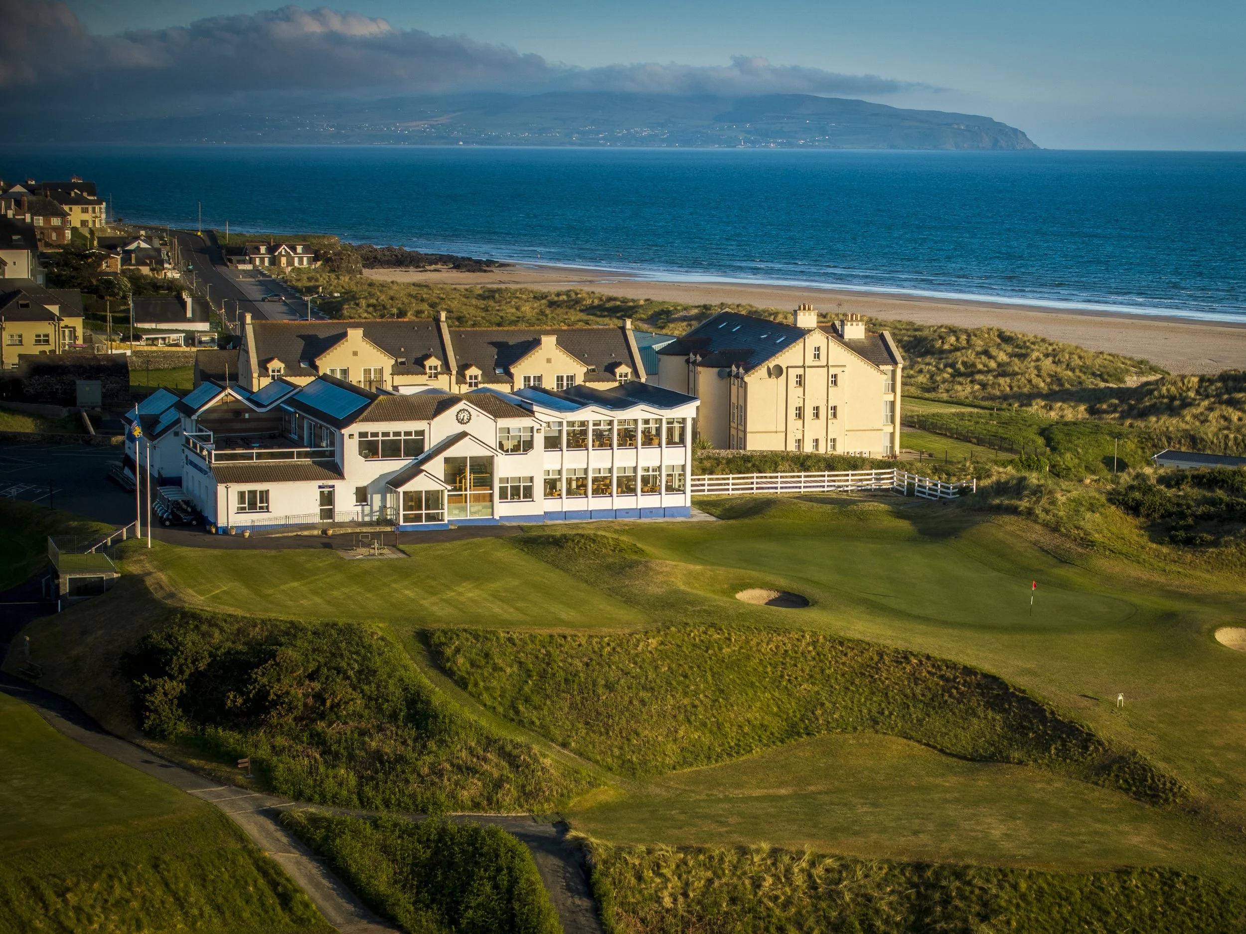 A scenic view of a coastal area with houses near the beach, a golf course with a green, and an expansive ocean with a distant landmass under a partly cloudy sky.