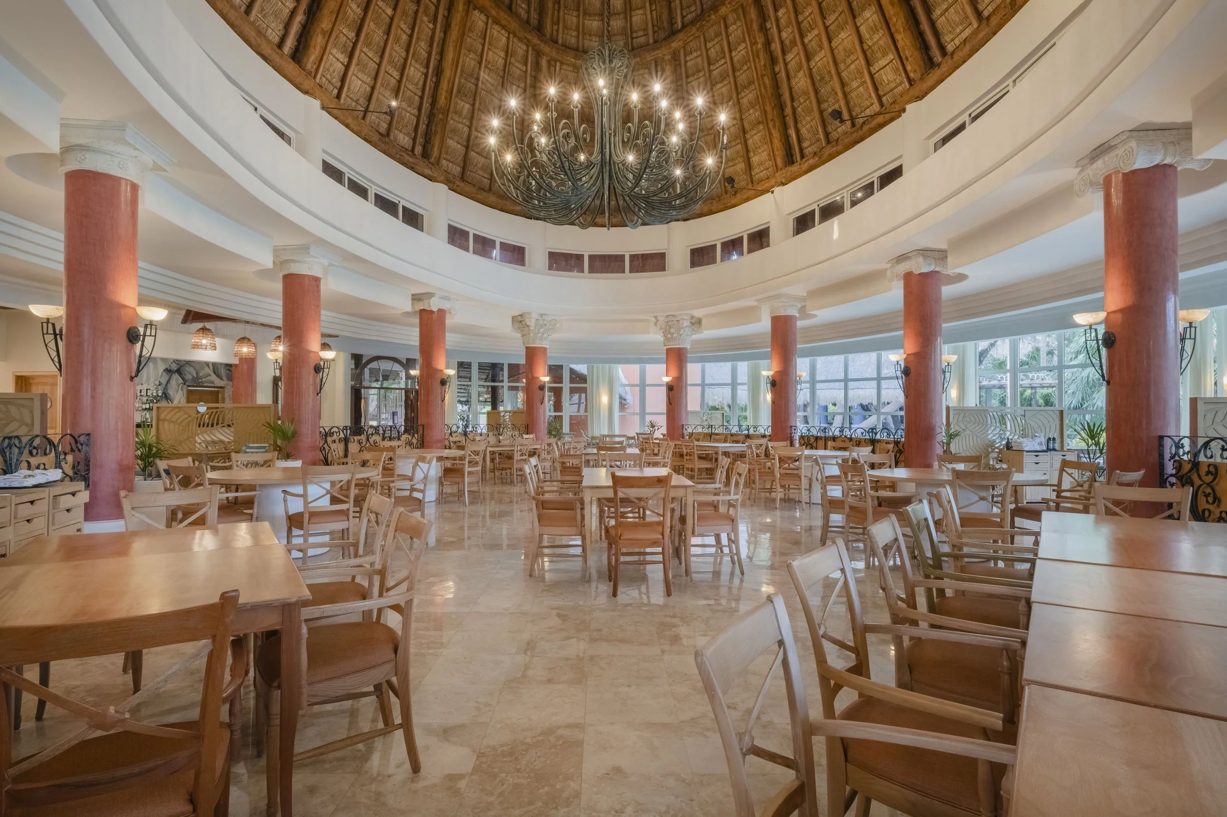 Interior of a spacious restaurant with wooden tables and chairs, tall red columns, large windows, and a high thatched ceiling with a central chandelier.