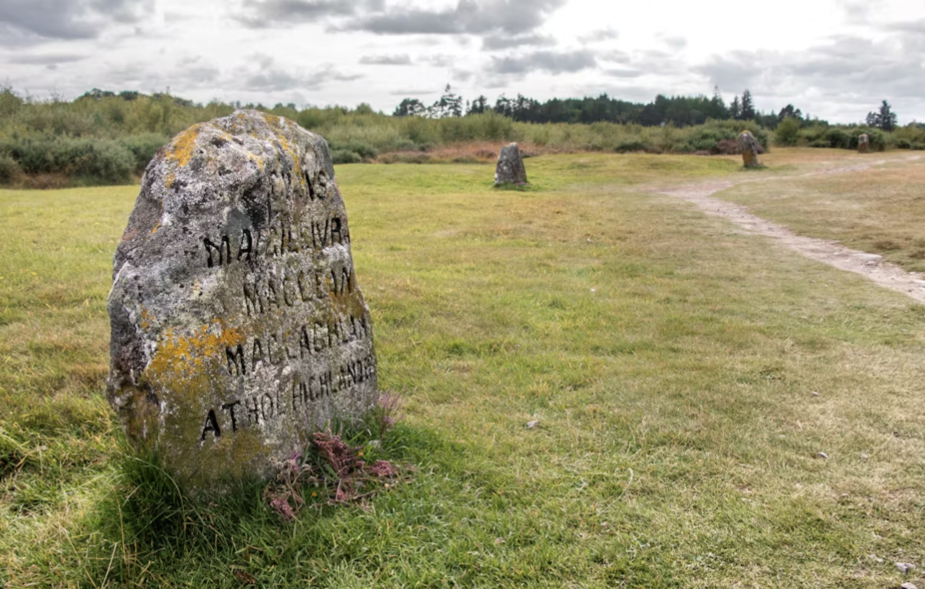 Ancient stone monument with engraved text in a grassy field with a dirt path, multiple weathered stones in the distance, and cloudy skies.