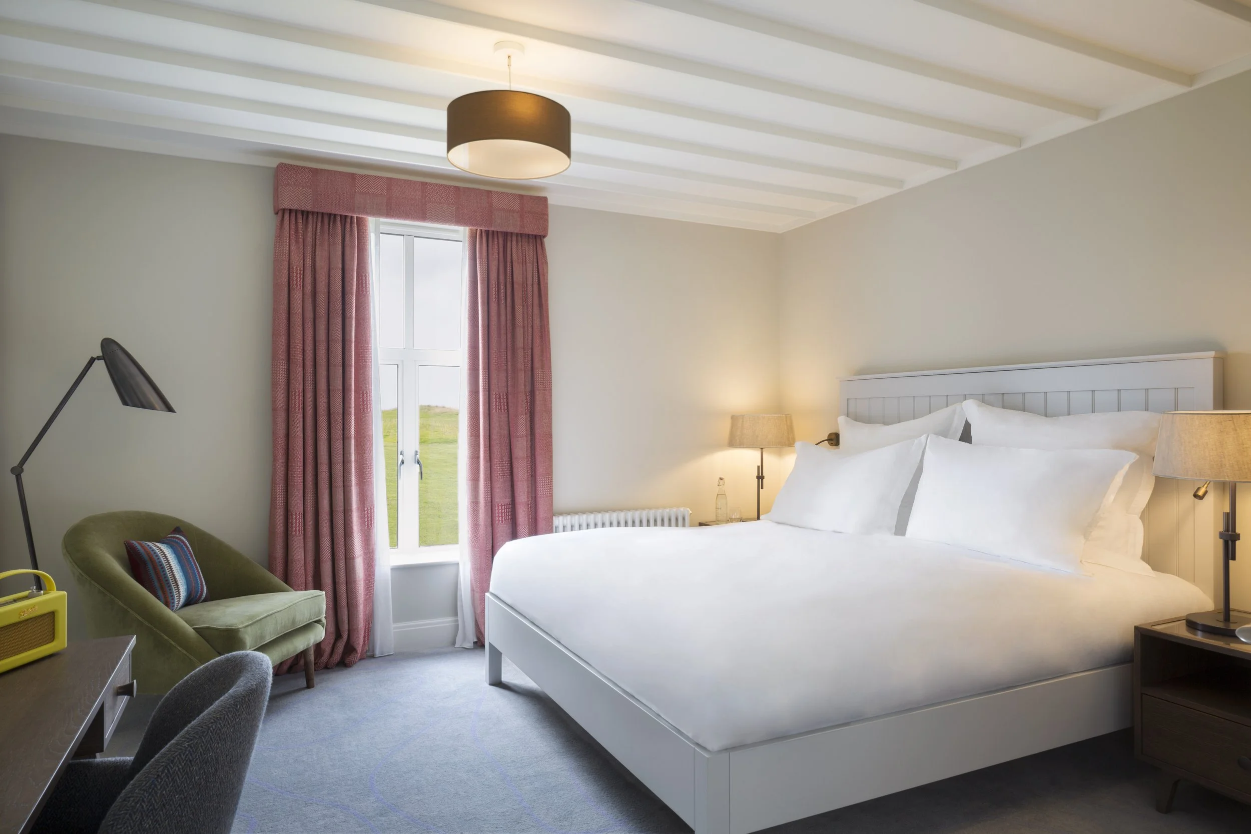A cozy hotel room with a large white bed, pink curtains, a green armchair with a striped pillow, and a window showing a grassy landscape outside.