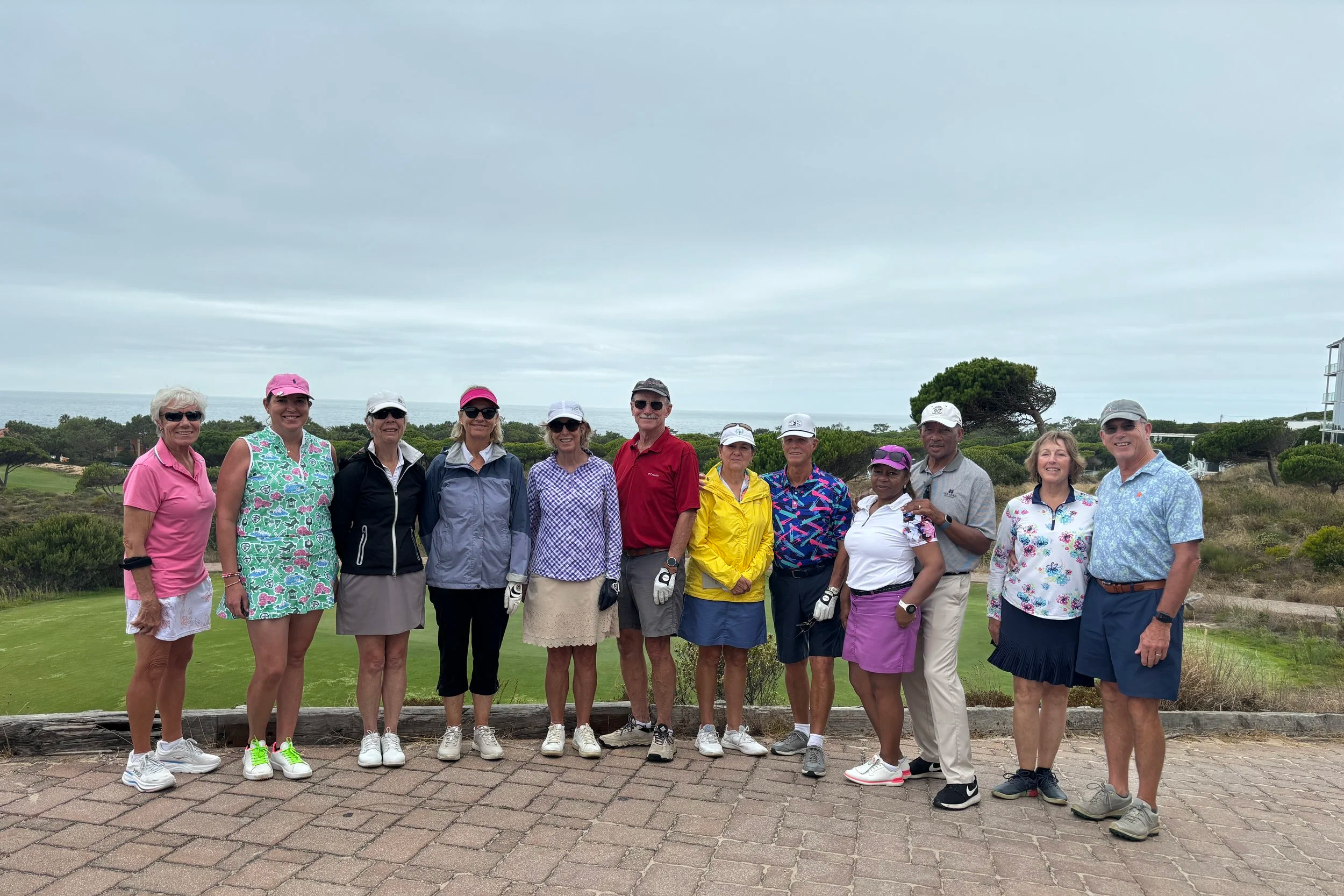 A group of twelve people dressed in golf attire standing outdoors on a golf course. Overcast sky in the background with trees and a building visible.