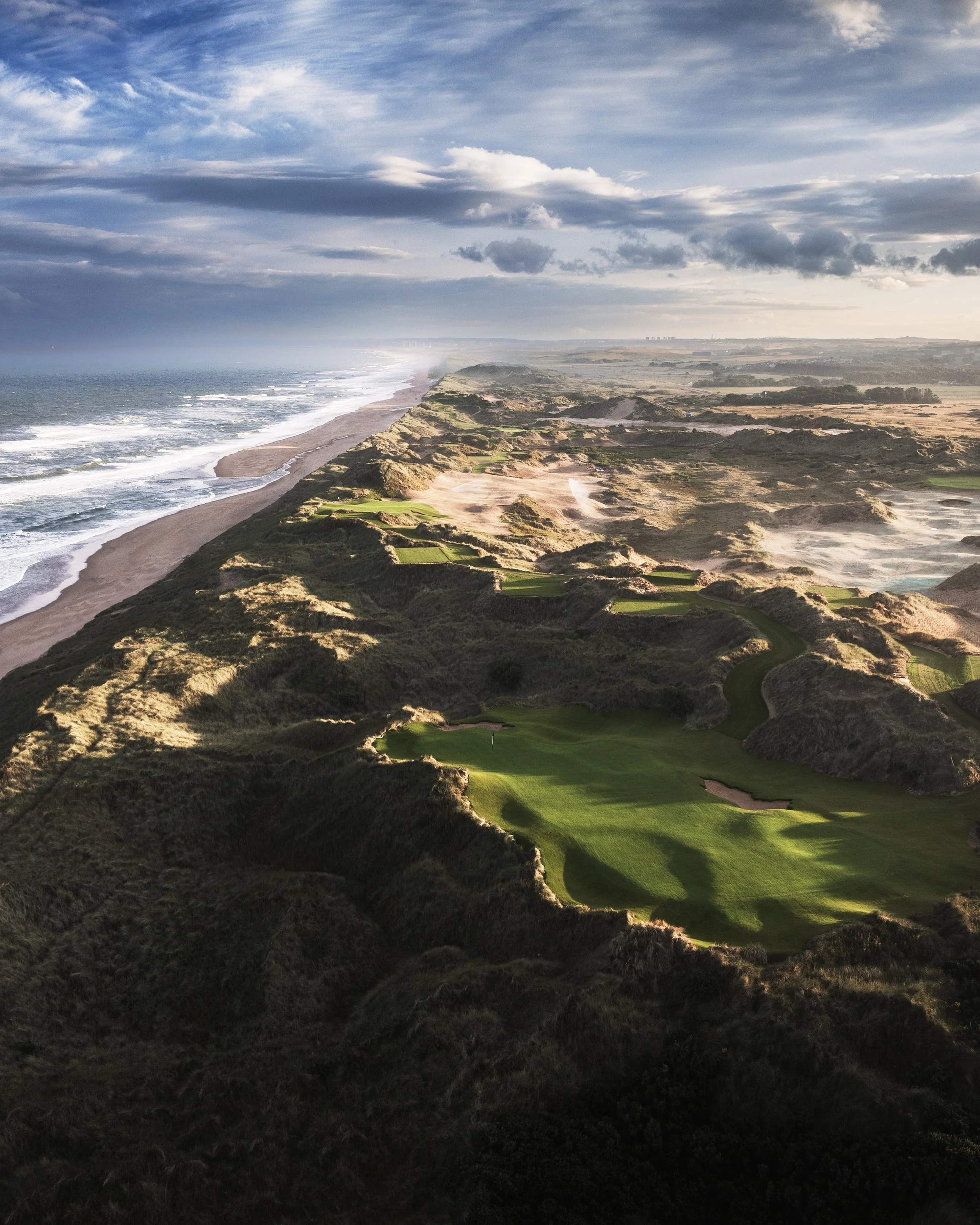 Aerial view of a coastal golf course with green fairways and sand traps, overlooking the ocean with sandy beaches and clouds in the sky.