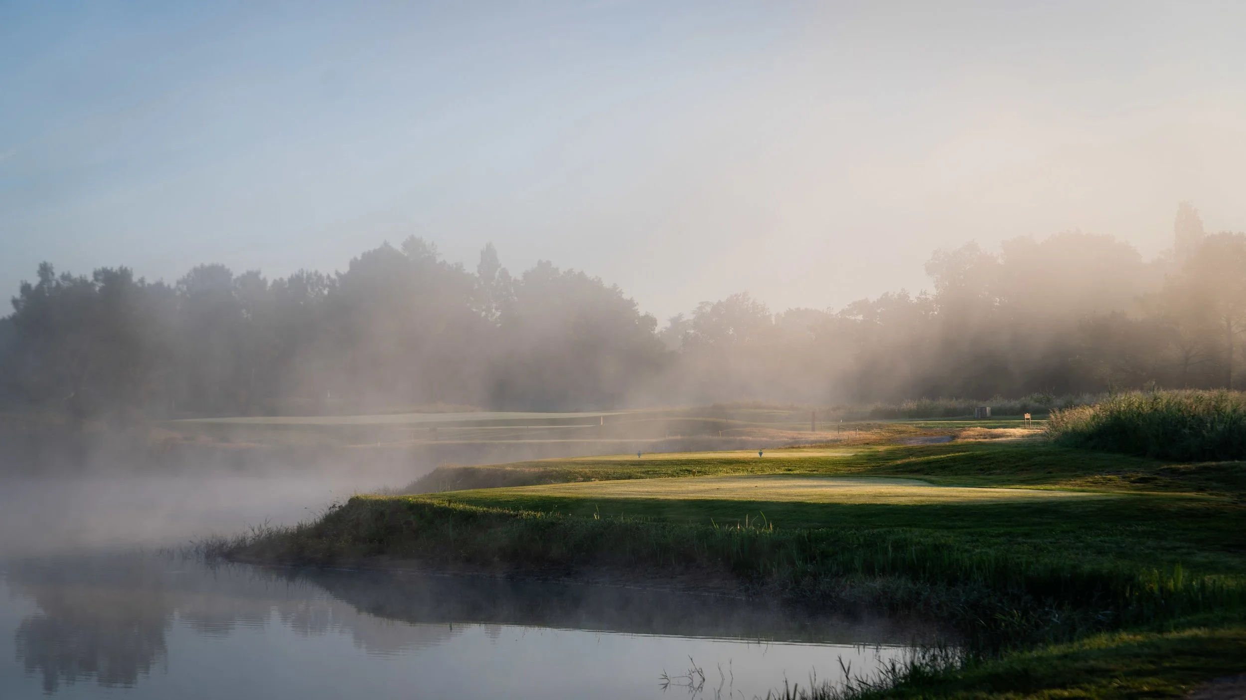 A serene misty morning view of a green fairway alongside a calm lake at Cabot Bordeaux, a breathtaking destination on our luxury escorted golf tours to France.