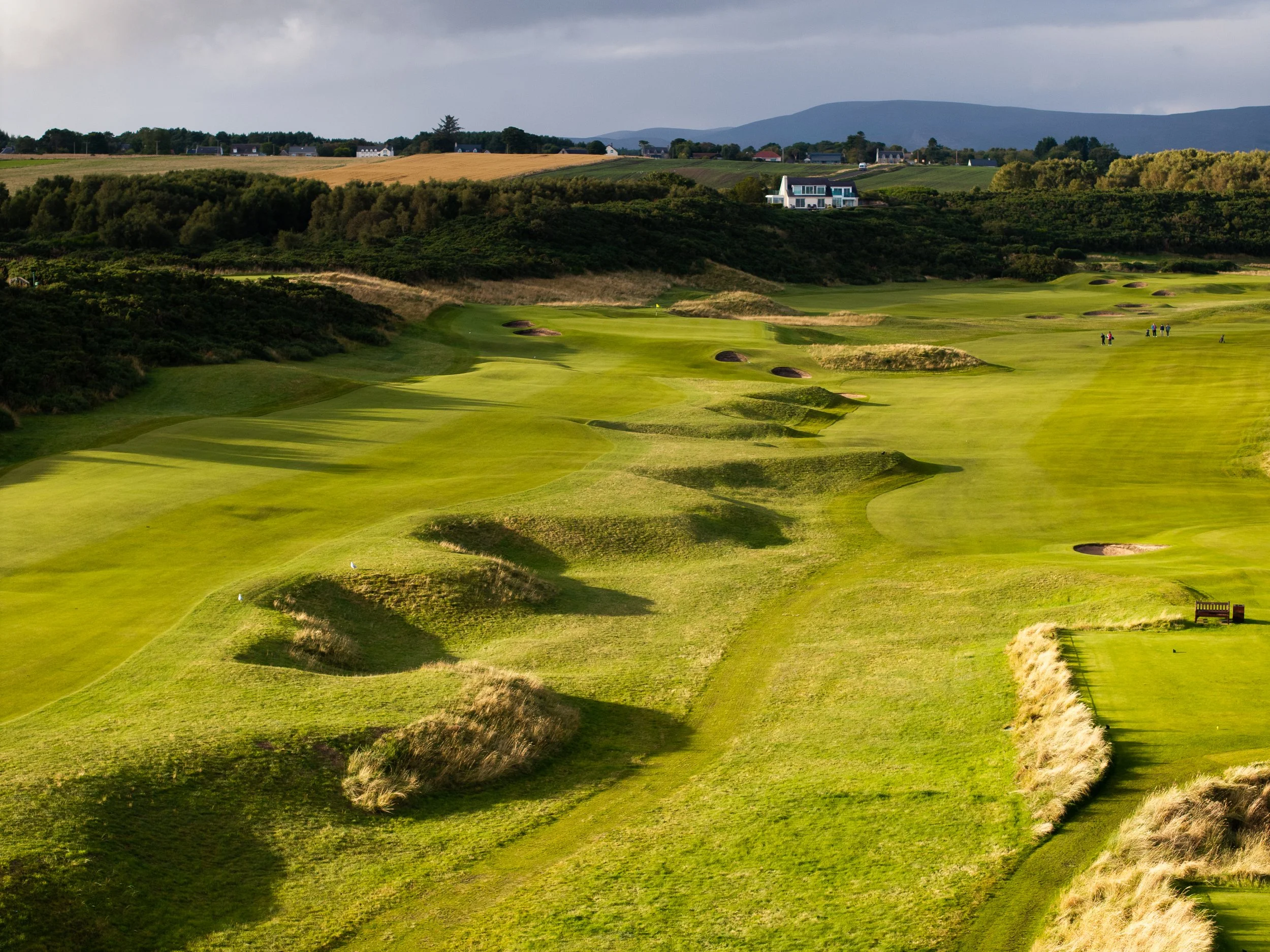 Green golf course with sand traps, rolling hills, and a group of people in the distance, with mountains and houses in the background.