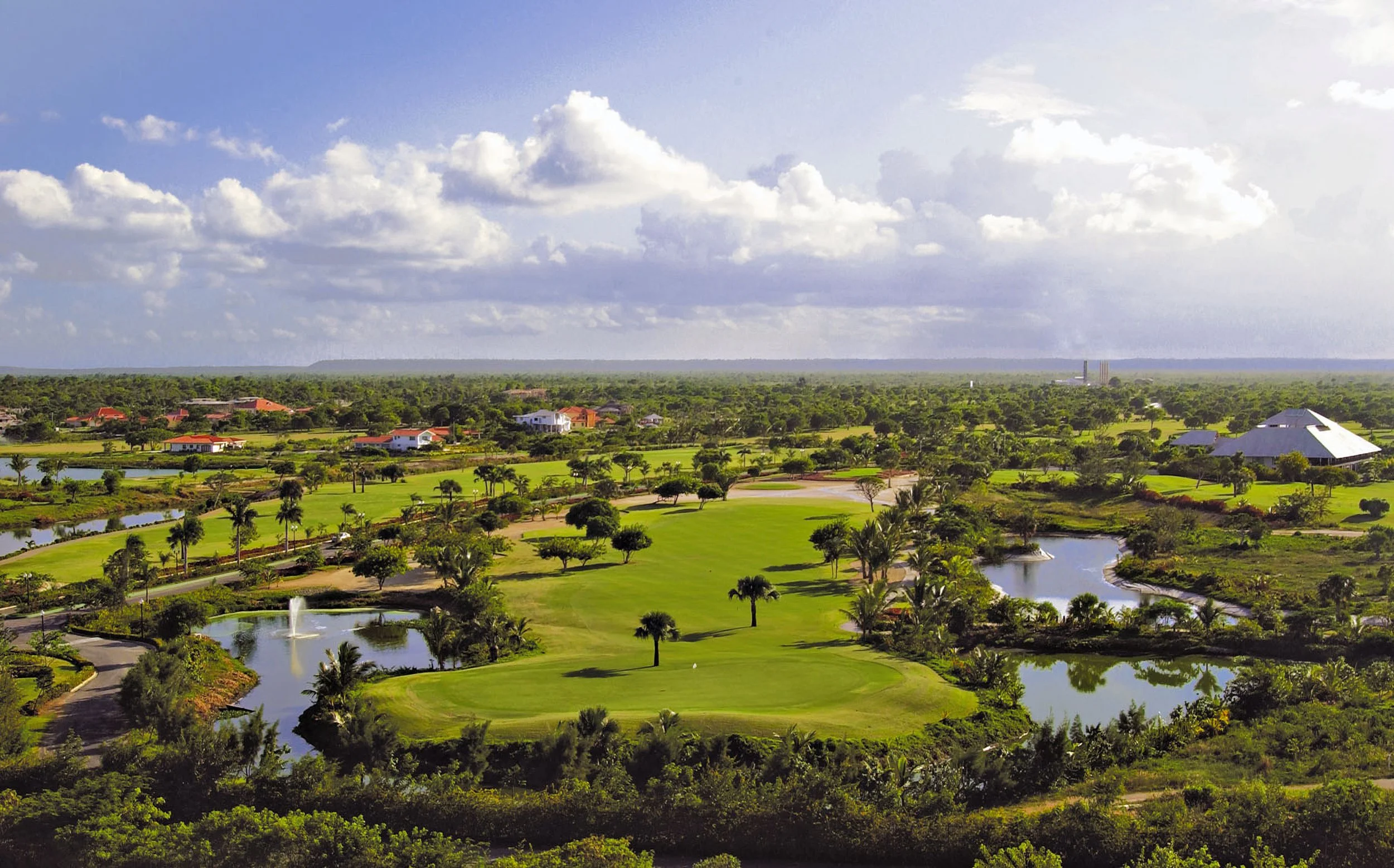A scenic view of a golf course with well-maintained greens, small lakes, palm trees, and a few buildings in the distance under a partly cloudy sky.