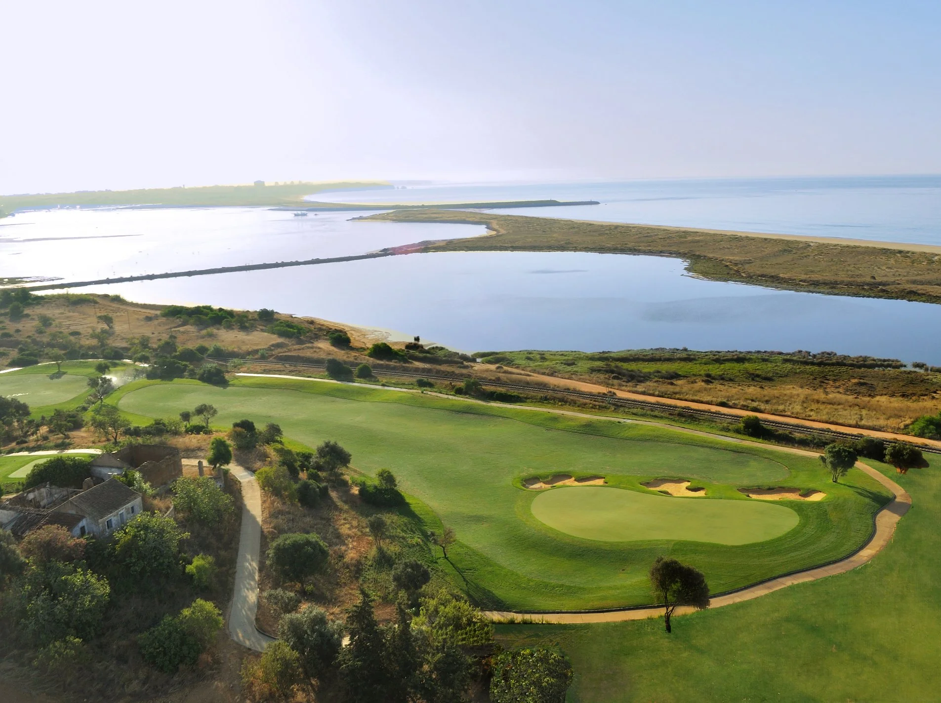 A golf course with green grass, sand bunkers, and trees near a river or lake, with water and distant land visible in the background.