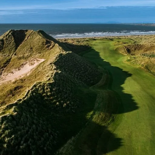 Aerial view of a coastal golf course, with sand dunes on the left and the ocean in the background.