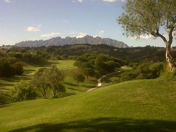A scenic golf course with green fairways, trees, and a winding path, with mountains in the background and a partly cloudy sky.