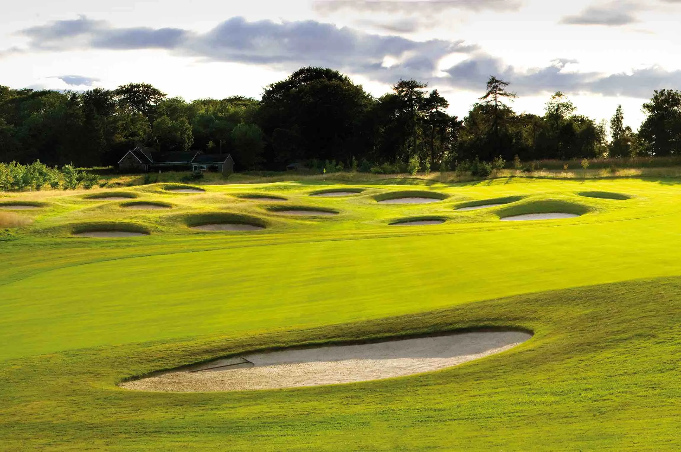 A golf course with green fairways and sand traps, surrounded by trees and a cloudy sky.
