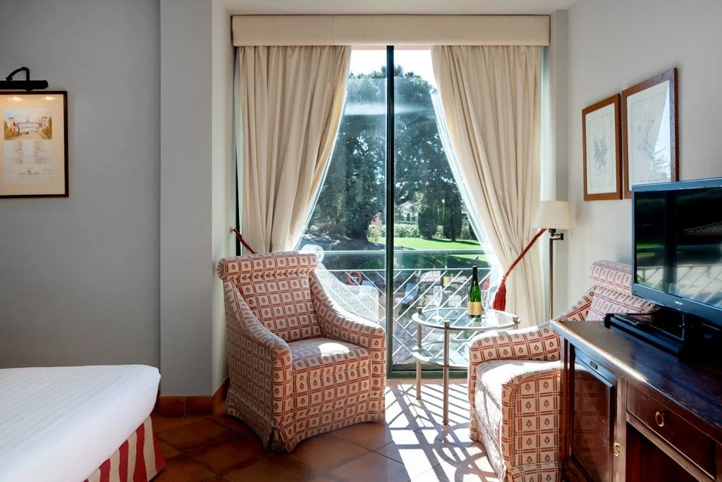 Hotel room with a sliding glass door opening to a balcony and view of trees outside. The room has two patterned armchairs, a small glass table with a bottle and glasses, a lamp, a TV on a wooden cabinet, and framed artwork on the walls.