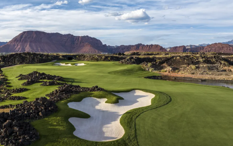 A scenic golf course with sand bunkers, green fairways, rocky terrain, and distant mountains under a partly cloudy sky.