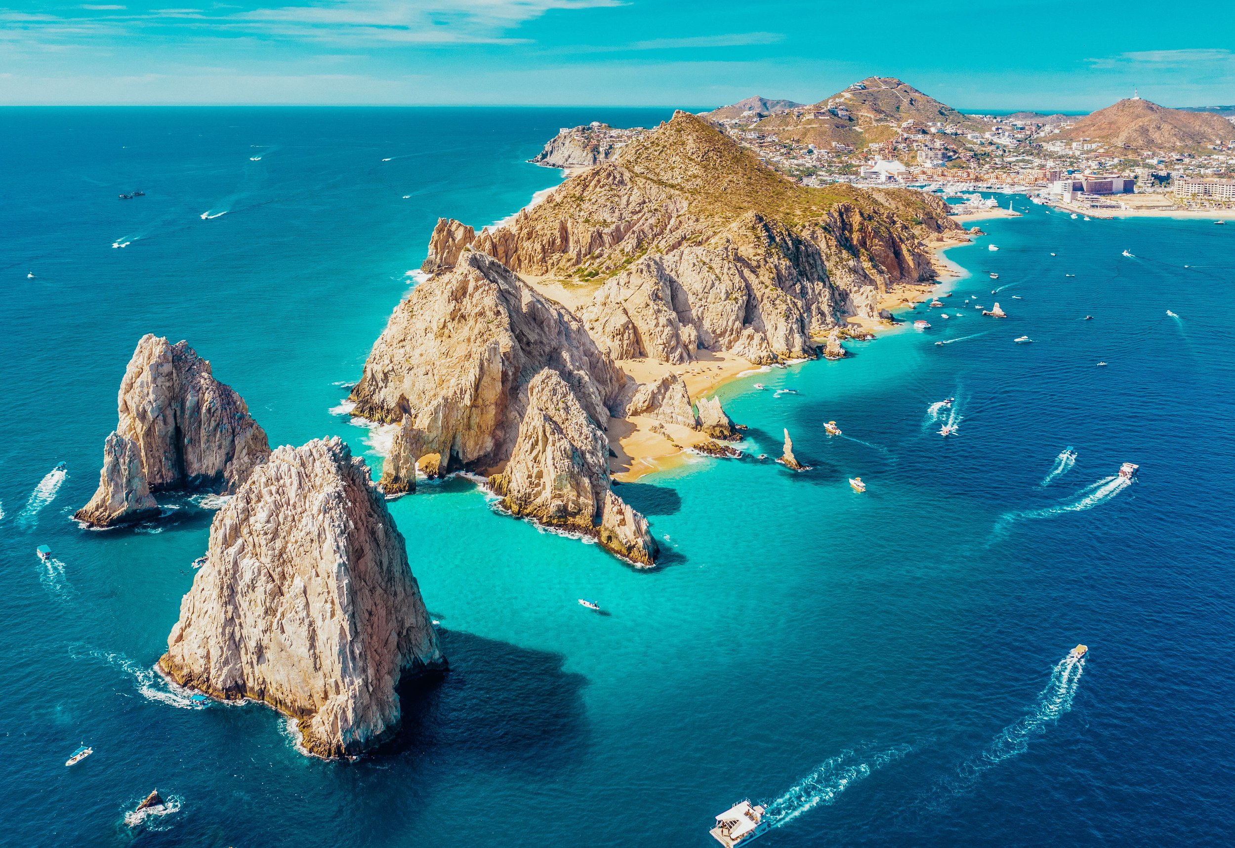 Aerial view of Land's End and the Rock forming the western tip of the Baja California Peninsula, with boats in the turquoise waters and the cityscape of Cabo San Lucas in the background.