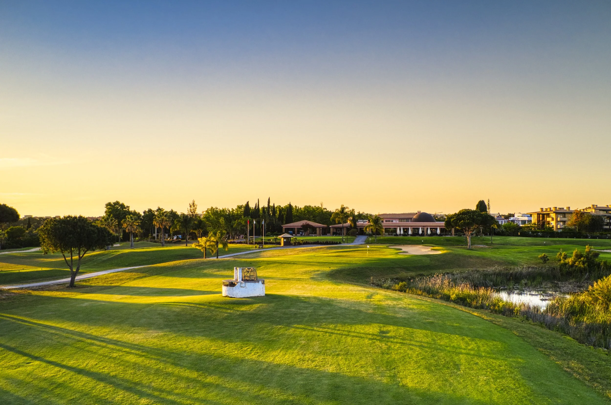 A golf course during sunset with green grass, trees, sand traps, a water hazard, and a clubhouse in the background.
