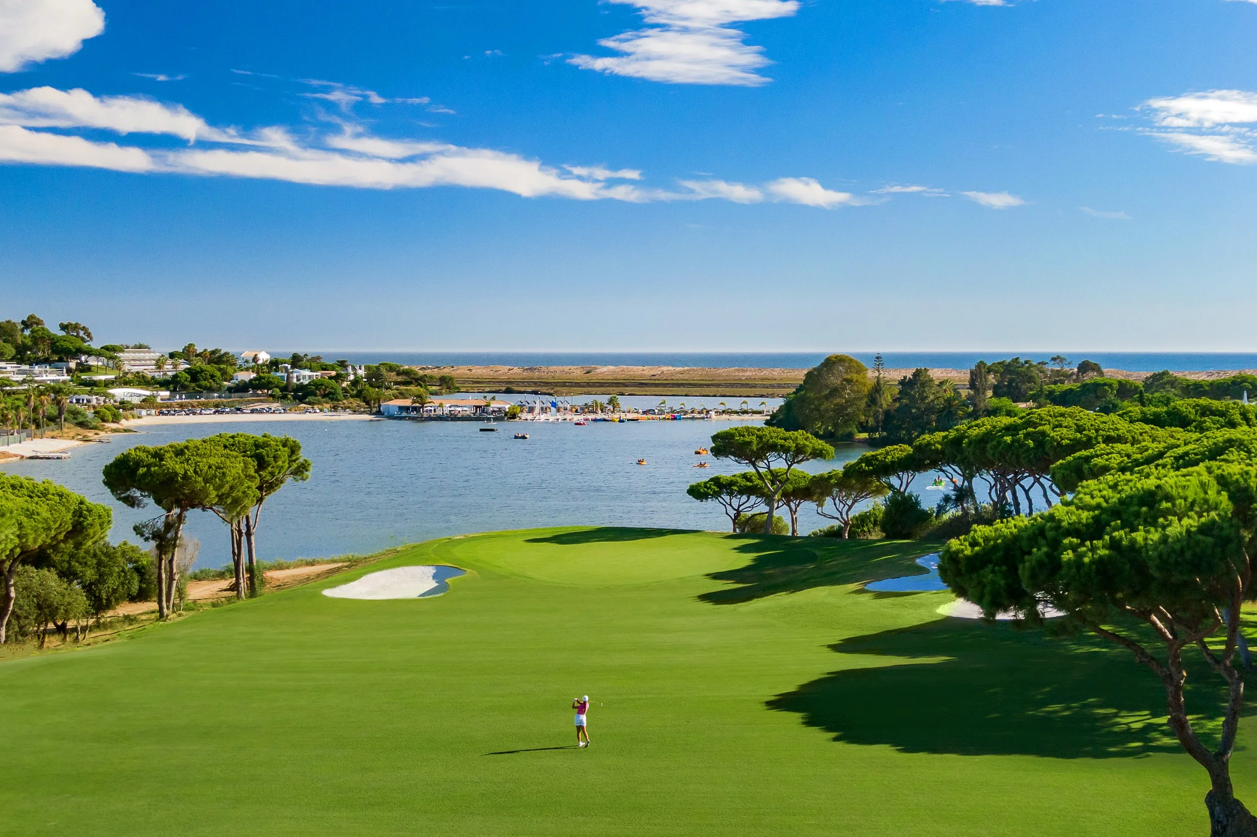 A golf course near a body of water with boats, and a cityscape in the background. The sky is blue with some clouds, and the course has trees and sand bunkers.