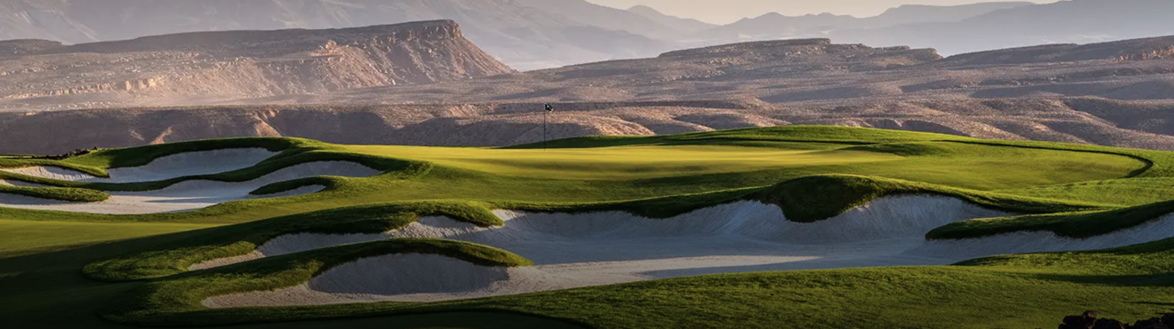 A golf course with sand traps and green fairways, set against a backdrop of mountains and a hazy sky.