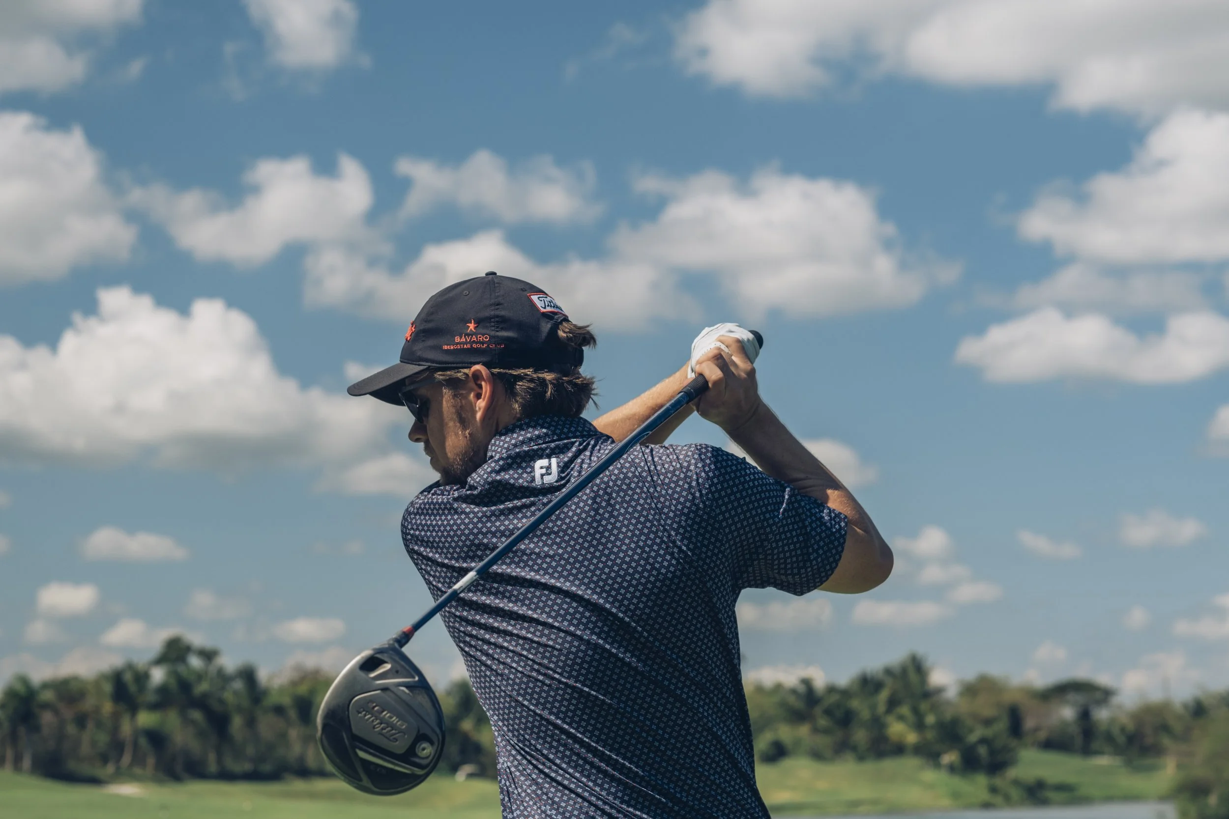 Man in a blue patterned shirt wearing a black cap with red logo, sunglasses, and white glove, swinging a golf club on a grassy course with trees and blue sky with clouds in the background.