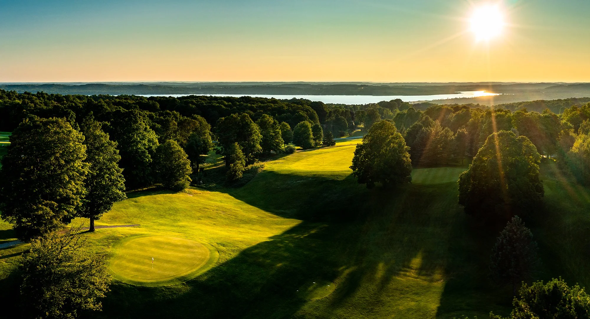 Sunset over a lush golf course with green fairways and trees, overlooking a body of water in the distance.