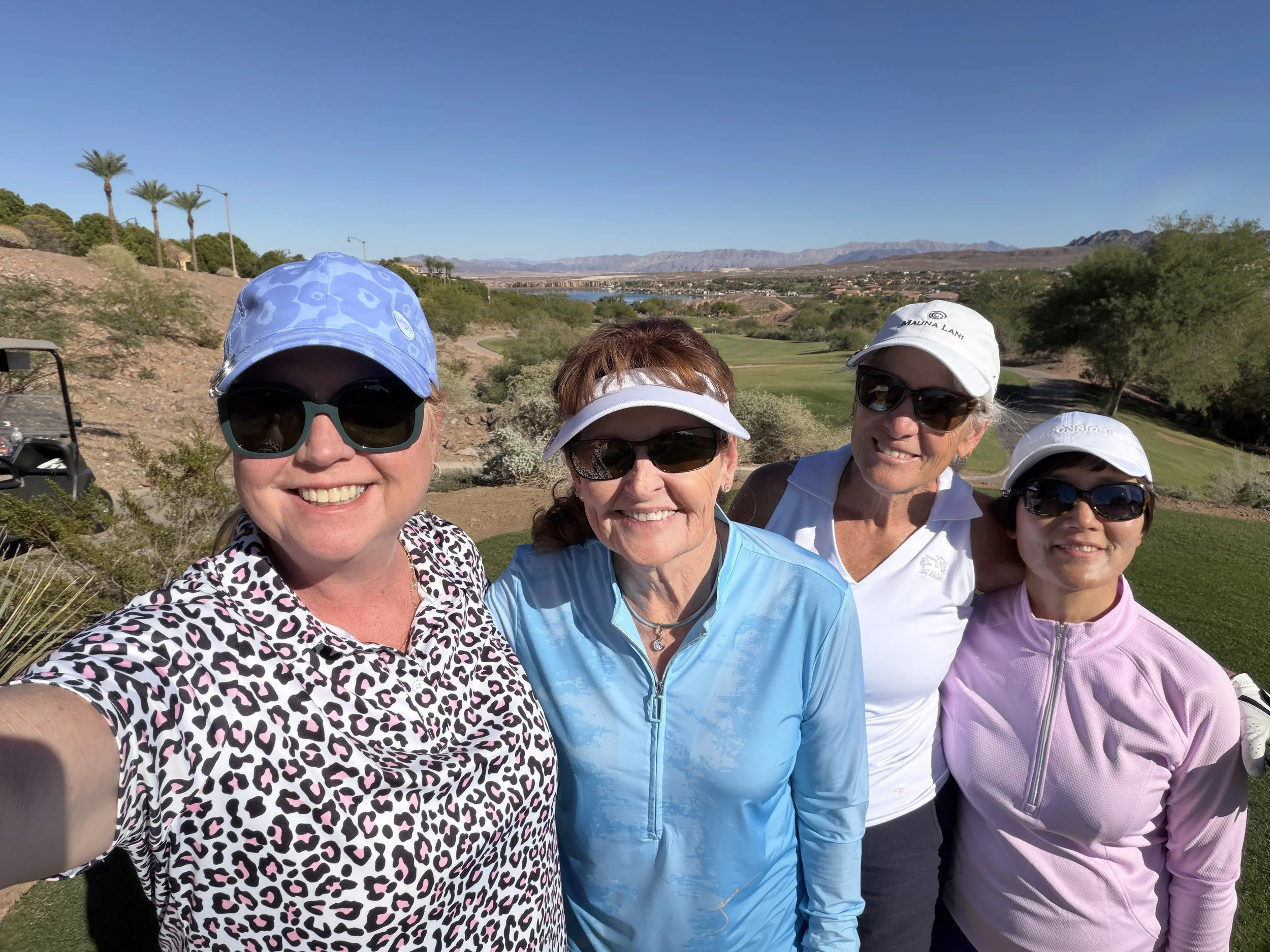 Four women smiling wearing golf hats and sunglasses on a golf course with desert landscape in the background.