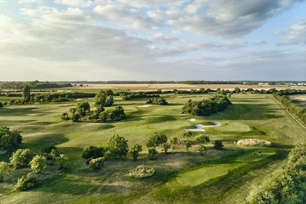 Aerial view of a golf course with multiple fairways, greens, sand bunkers, trees, and open landscape under a partly cloudy sky.