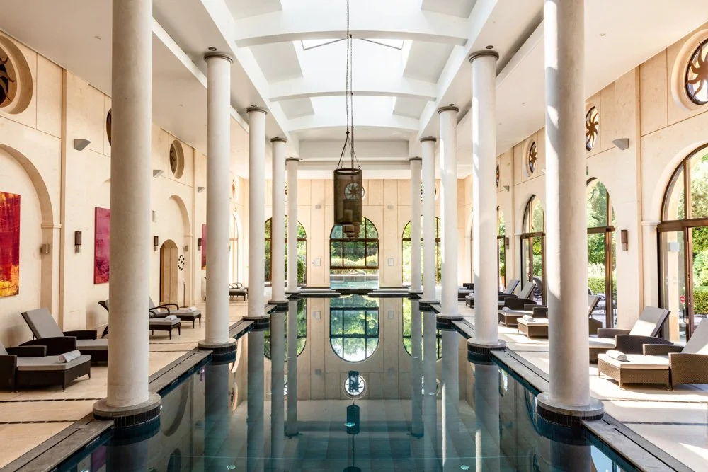 A serene indoor swimming pool framed by elegant white columns, large arched windows, and comfortable lounge chairs at the Terre Blanche Hotel spa, offering the perfect relaxing getaway during our luxury ladies' golf retreats in the French Riviera.