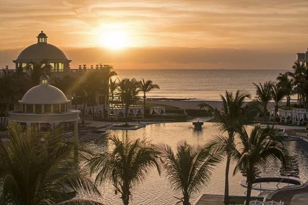 A luxurious resort pool area at sunset with palm trees, gazebos, and the ocean in the background.