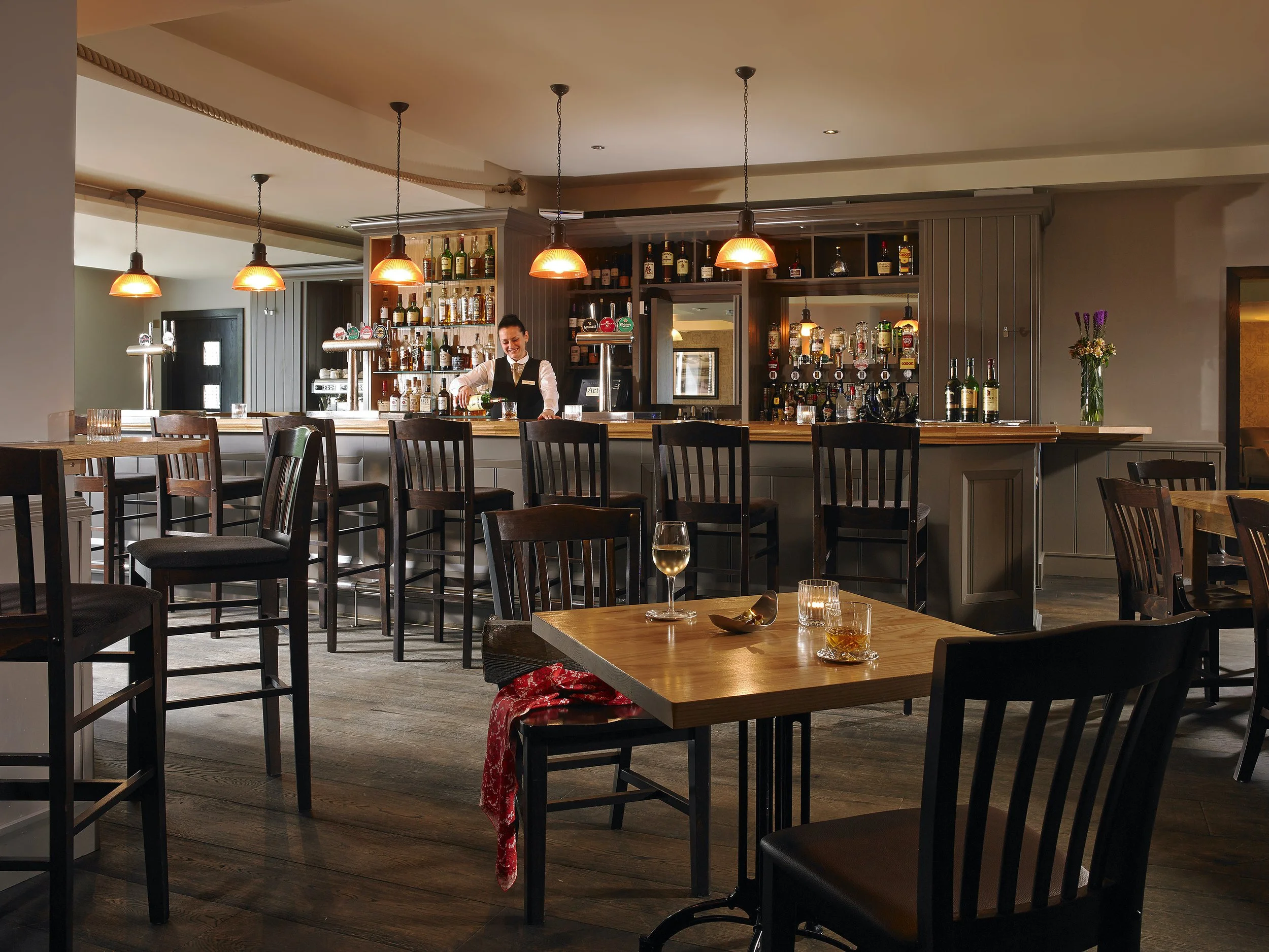 Indoor bar and restaurant with a bartender preparing drinks behind the bar. Several tables and chairs are arranged, some with glasses of wine and drinks on them. The atmosphere is cozy and warm with soft lighting.