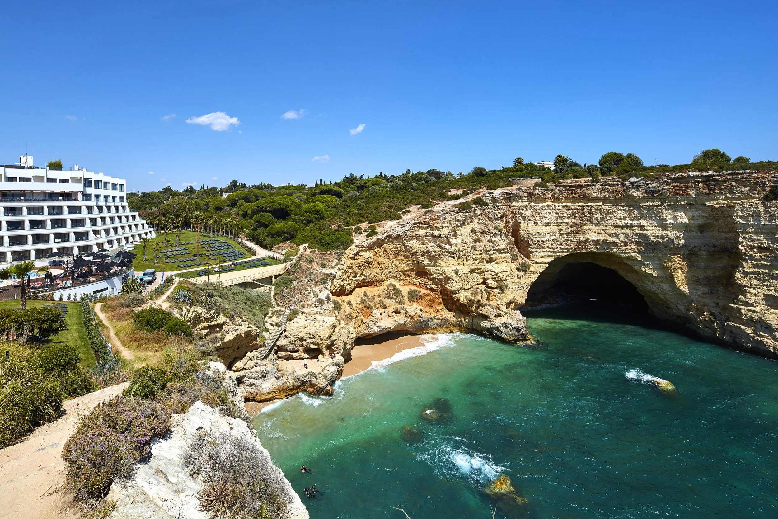 Beach with cliffs and a large sea cave, clear turquoise water, and a hotel with landscaped gardens nearby, under a blue sky with some clouds.