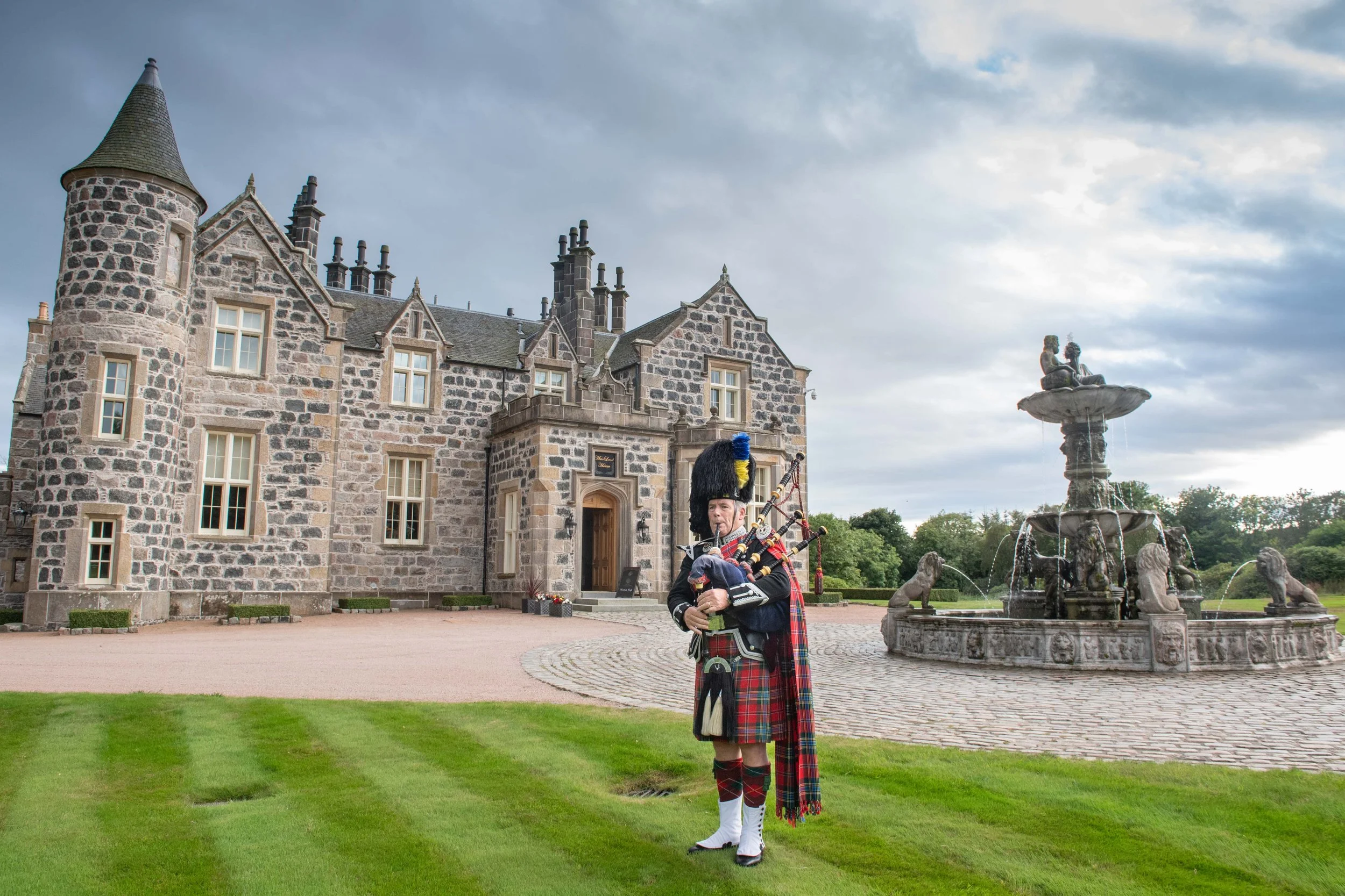 A man dressed in traditional Scottish kilt and uniform, playing bagpipes in front of a historic stone castle with a fountain in the courtyard.