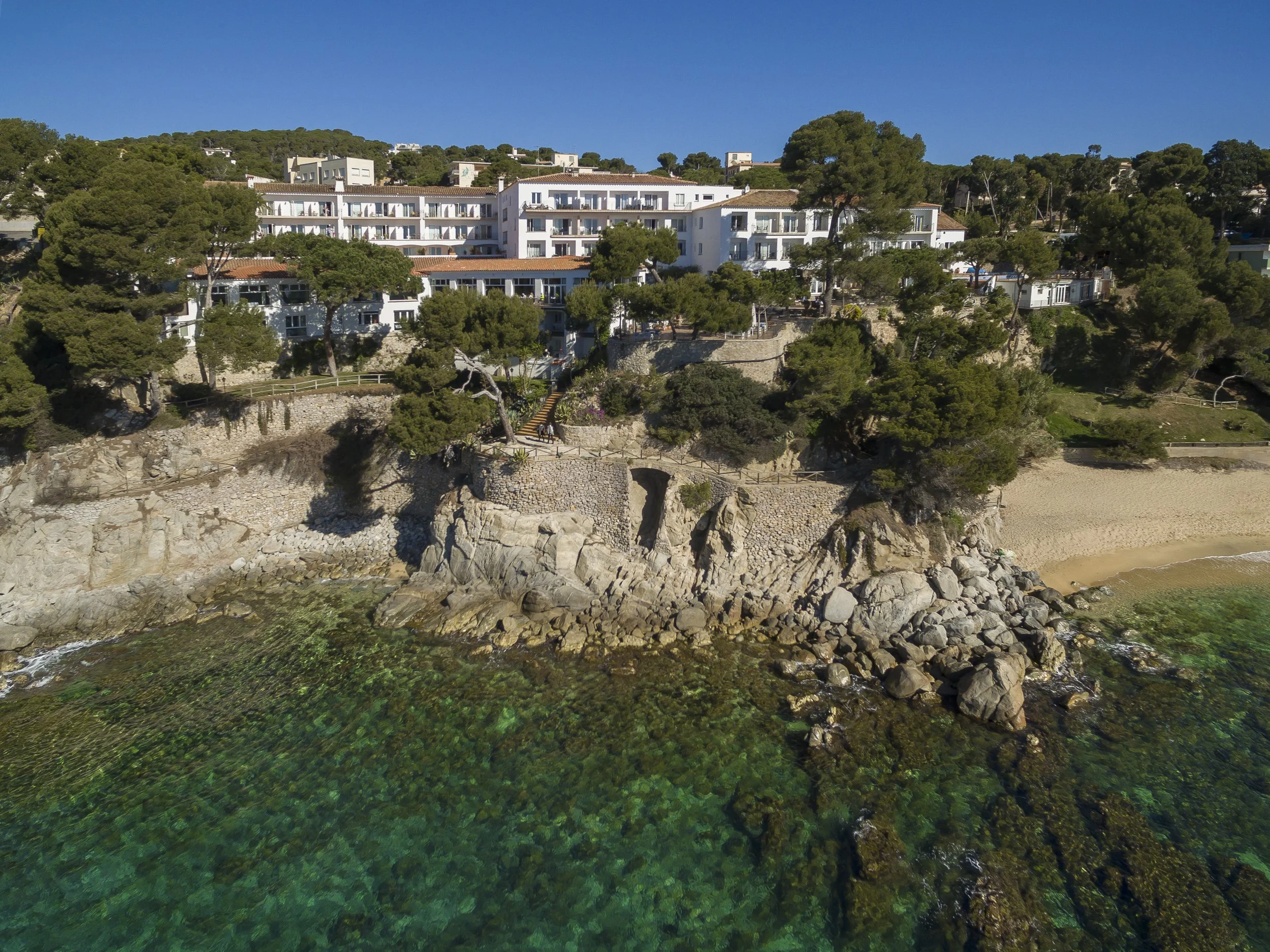 Aerial view of a coastal hillside with white residential buildings, trees, and a rocky beach, under a clear blue sky.
