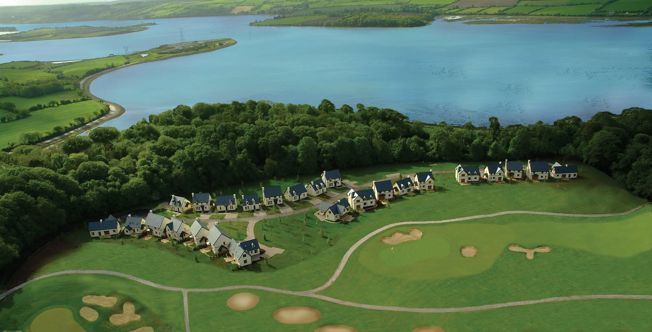 A residential golf course development with houses along a winding road, surrounded by trees and green fairways with bunkers, near a large body of water.