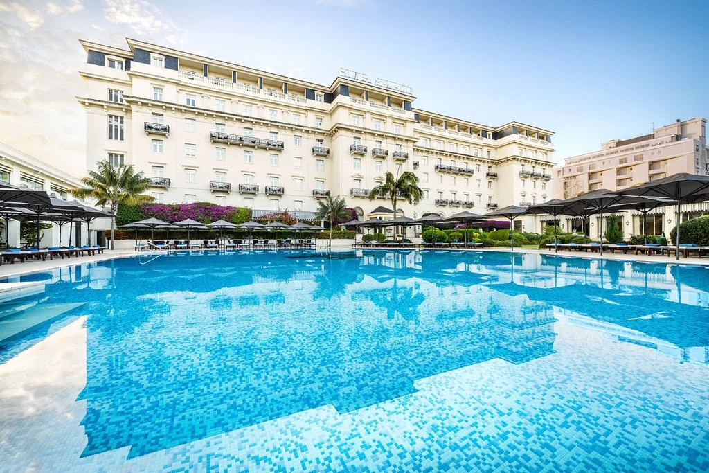 A luxurious hotel pool area with a large rectangular pool, surrounded by sun loungers with umbrellas, tall palm trees, and a white multi-story hotel building in the background under a partly cloudy sky.