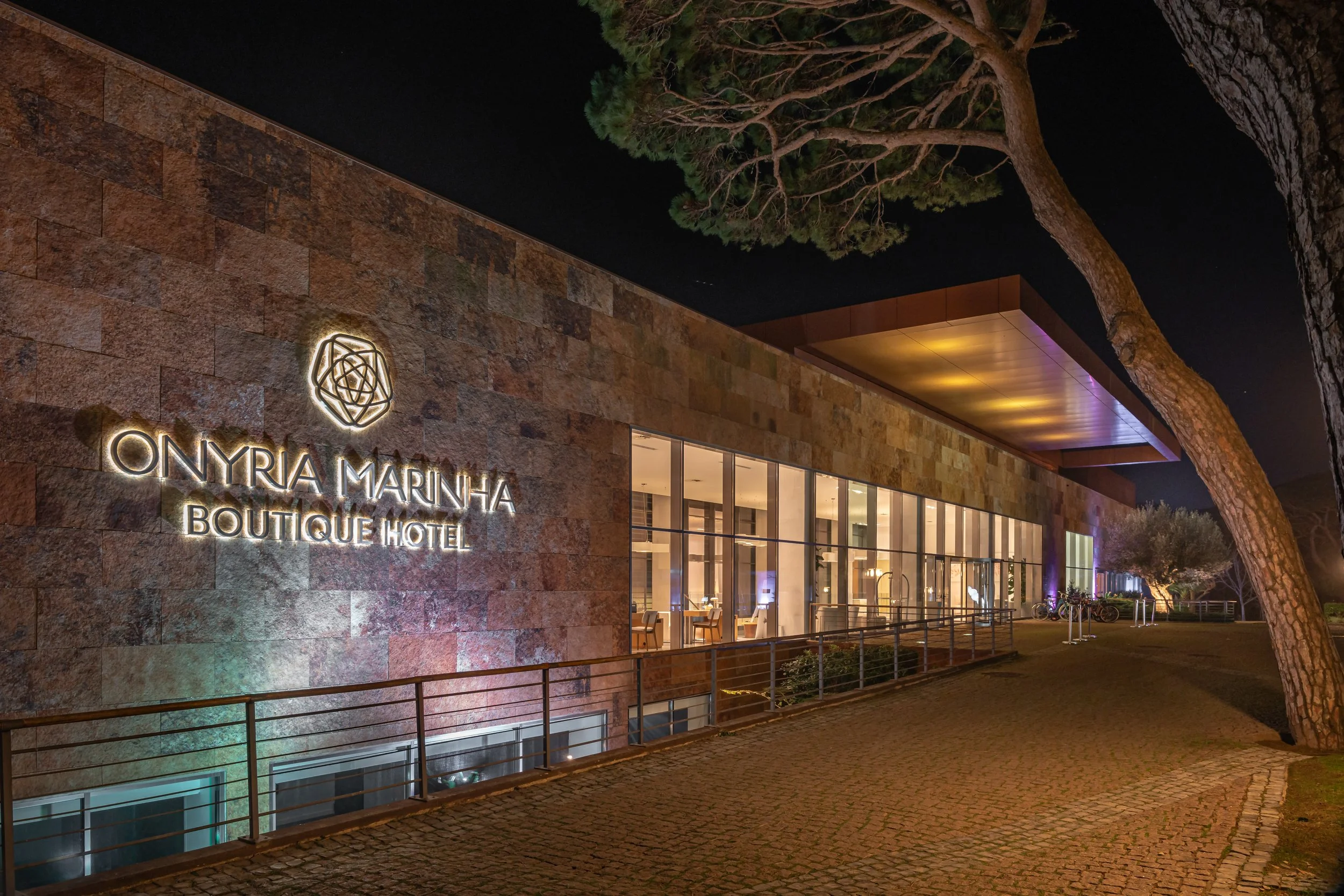 Night view of Onyria Marinha Boutique Hotel exterior with illuminated sign on a stone wall, large glass windows revealing the interior, and a tall tree in the foreground.