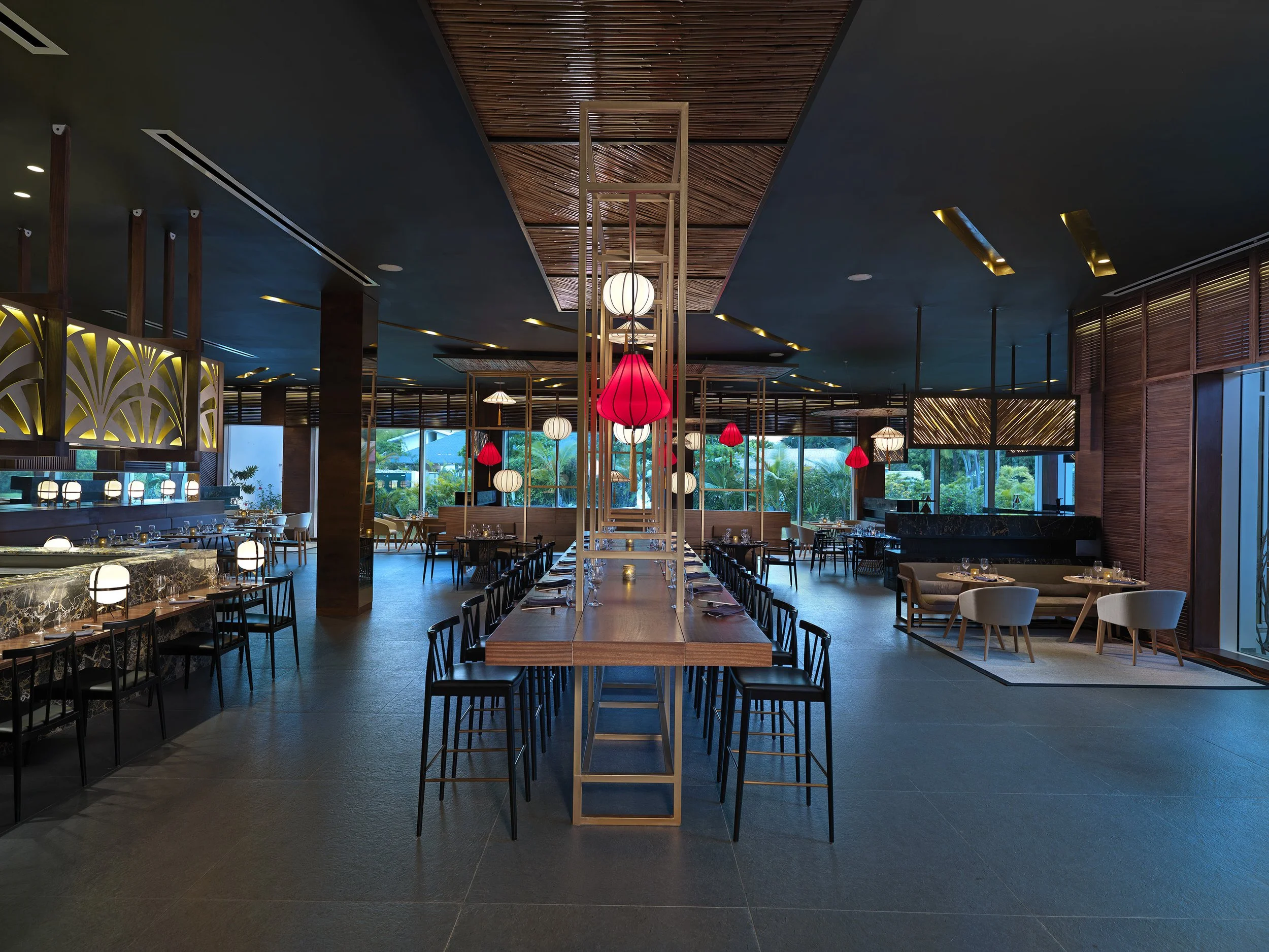 Interior of a modern restaurant with a high ceiling, wooden accents, and large windows showing greenery outside. There are hanging red and white lantern-style lights, a marble-topped bar area on the left, and tables with chairs arranged throughout th