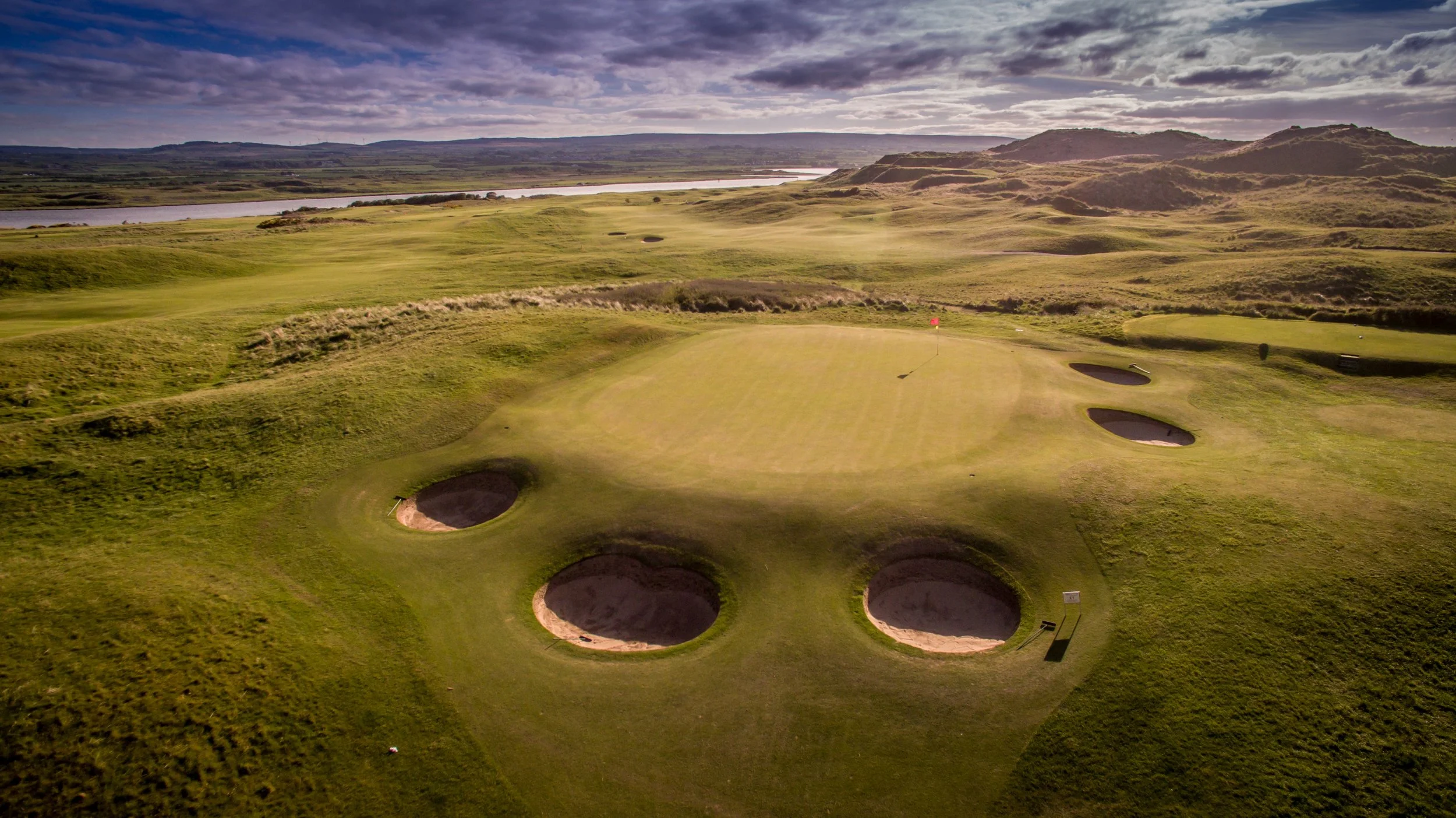 A golf course with multiple sand bunkers surrounding the green, set in a landscape of rolling hills and a river in the background, under a partly cloudy sky.