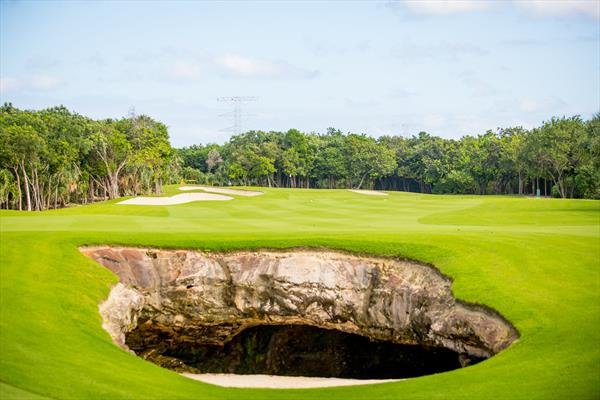 A golf course with a large, natural rock formation and a sand bunker, surrounded by green grass and trees under a partly cloudy sky.