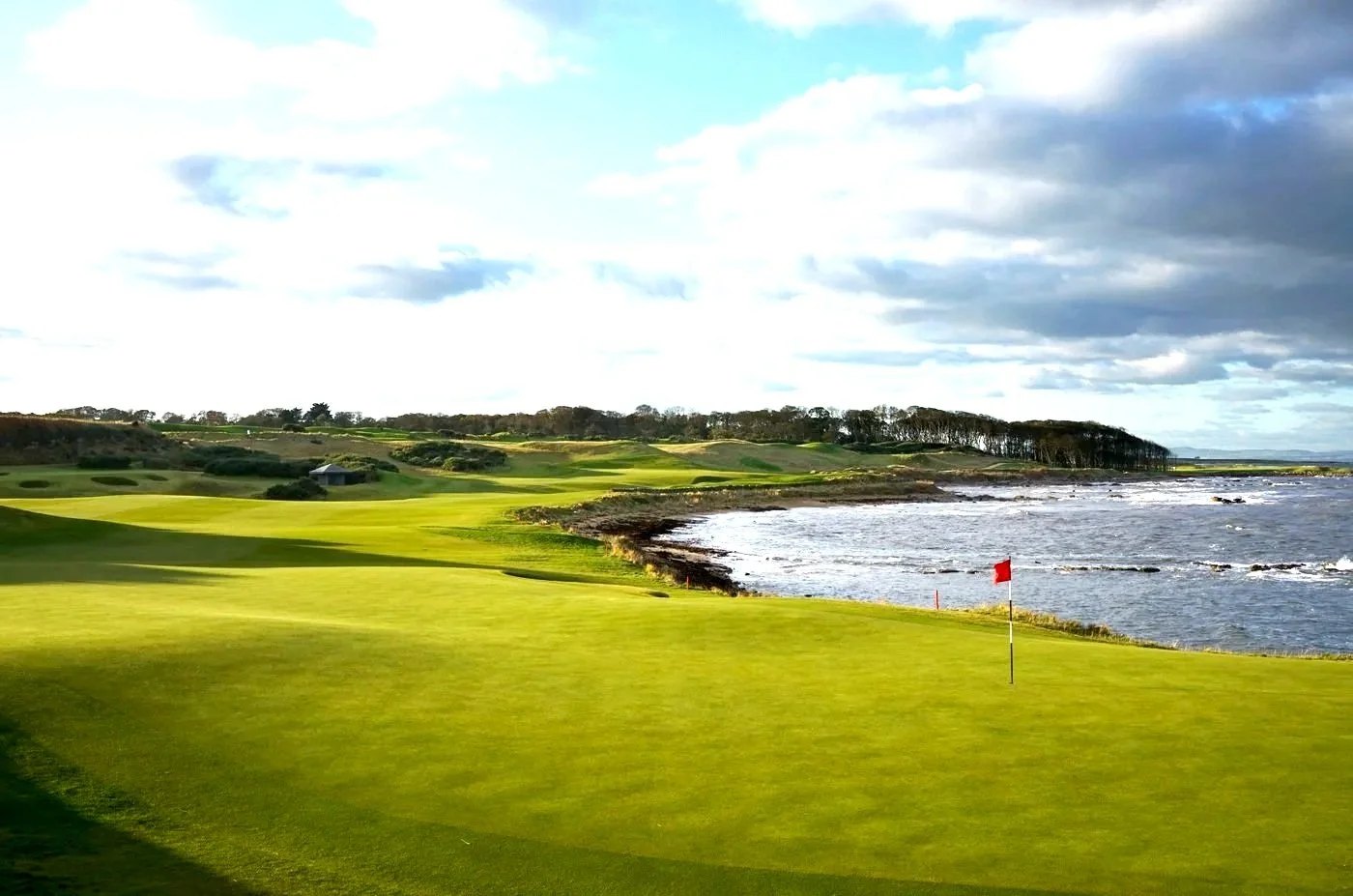 A scenic coastal golf course with green fairways and a flagstick near the water, under a partly cloudy sky.