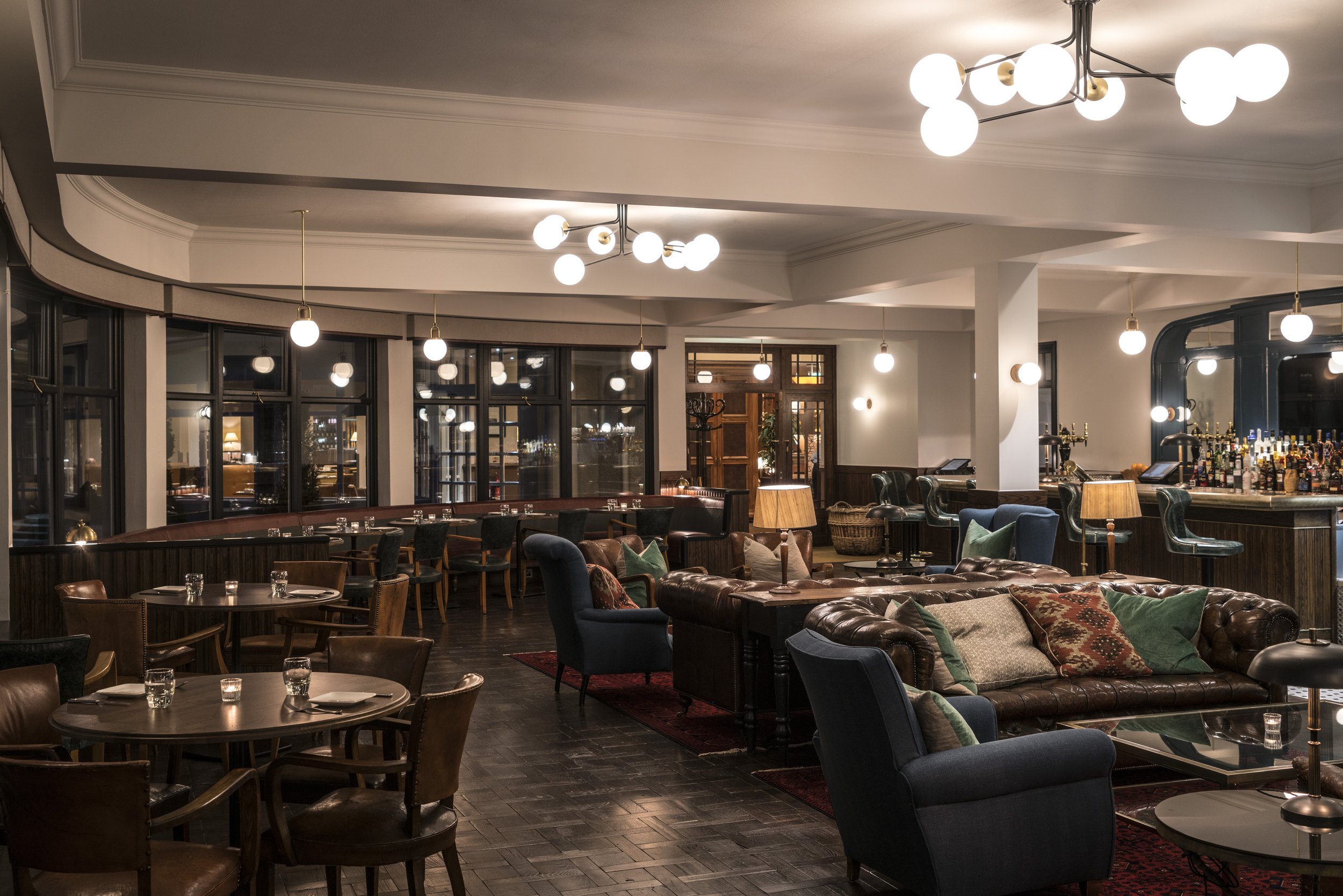 Interior of an elegant lounge bar with leather and upholstered seating, wooden tables, modern light fixtures, and a well-stocked bar area in the background.