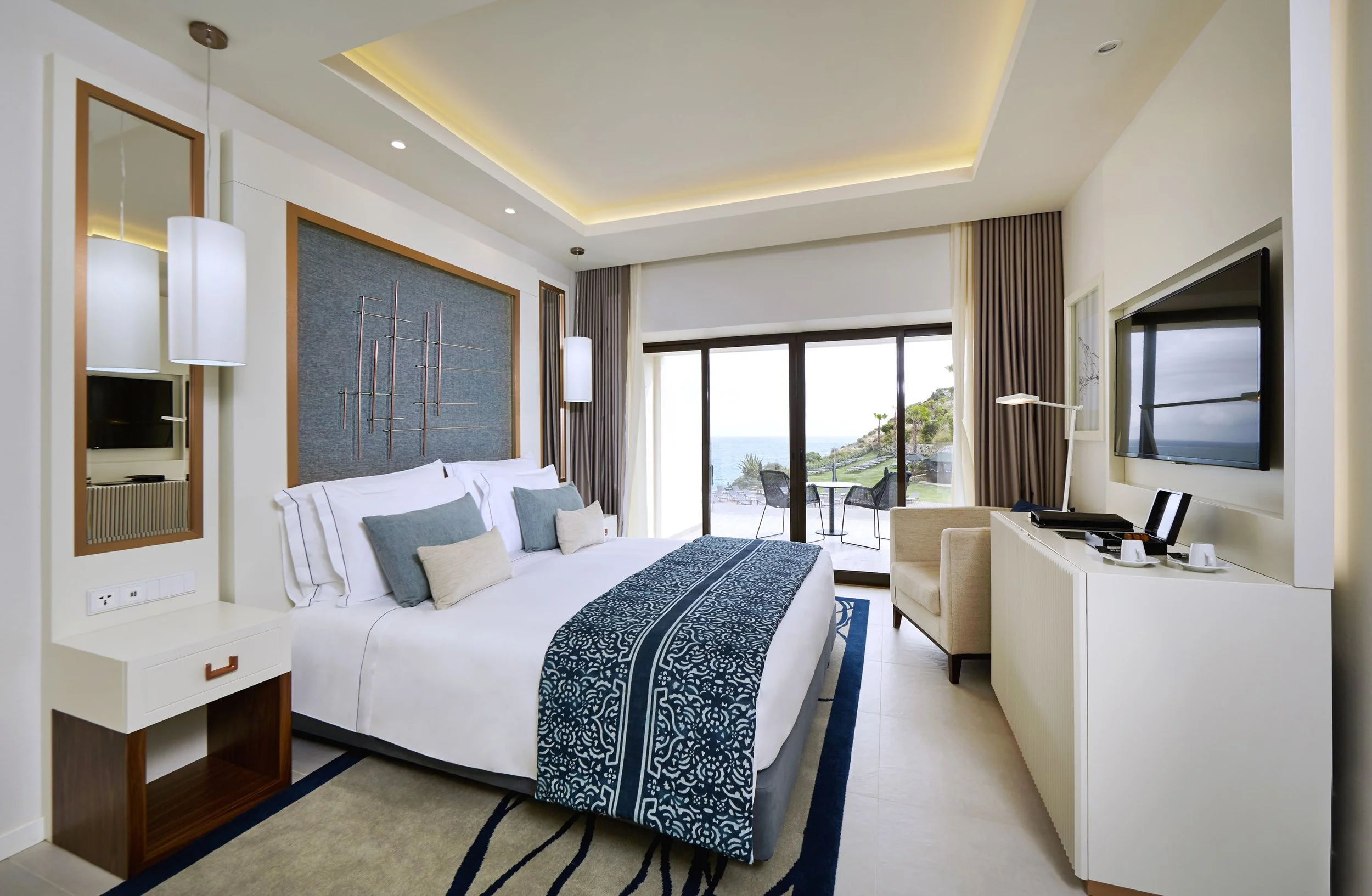 Modern hotel room with a king-sized bed, decorative pillows, blue patterned bed runner, sliding glass door leading to a balcony with ocean view, mounted flat-screen TV, white dresser, sitting chair, and a scenic landscape outside.