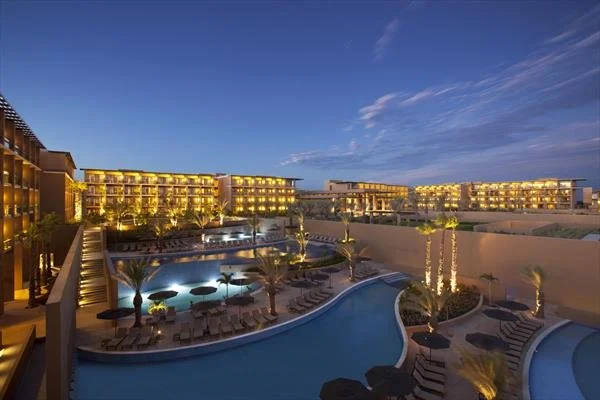 Night view of a resort with illuminated buildings, a large swimming pool, palm trees, and poolside lounge chairs.