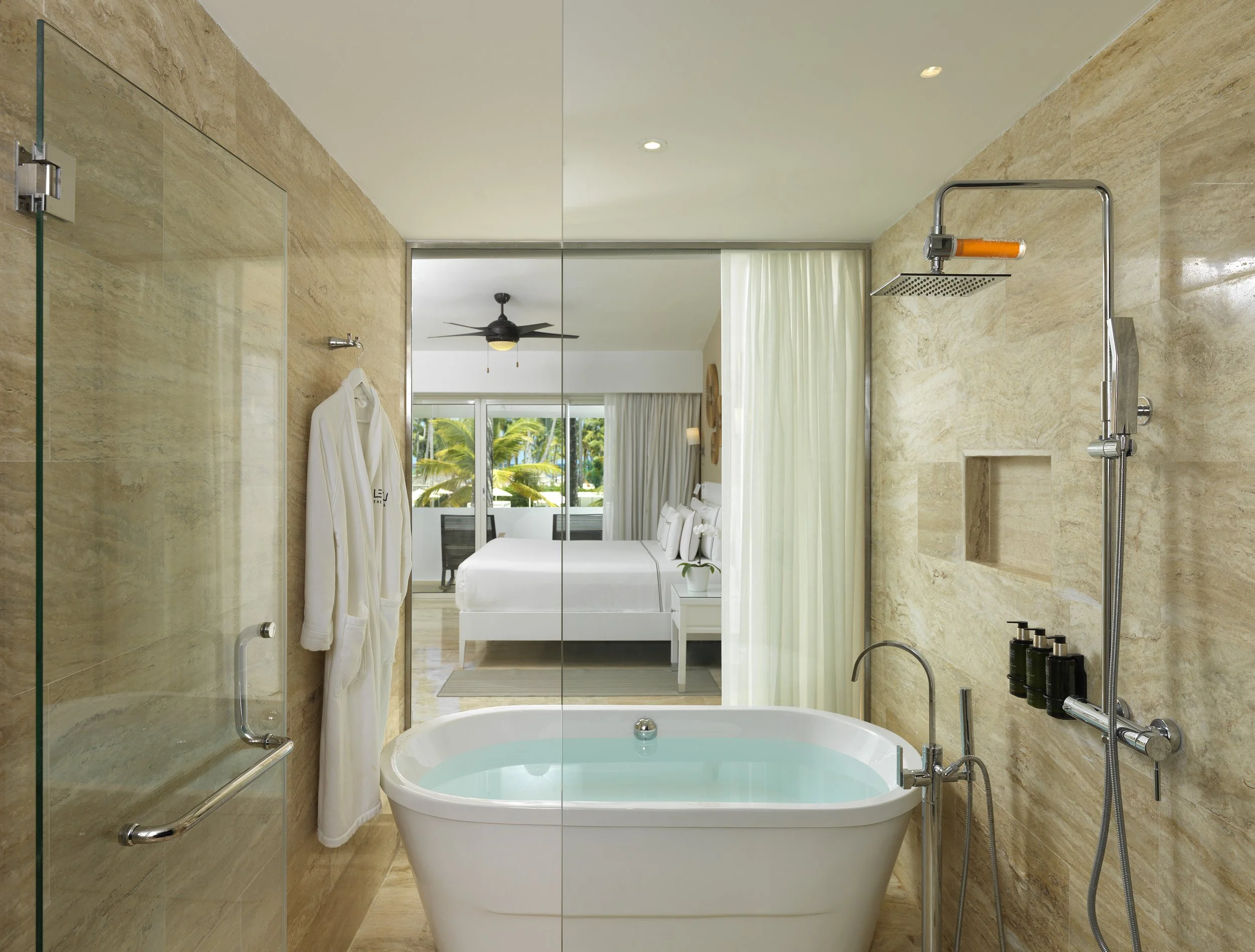 Modern bathroom with a bathtub filled with water, a showerhead, a robe, and a view of a bedroom with large windows and tropical trees outside.