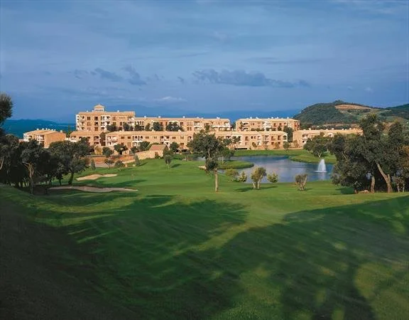 Golf course with green fairways, water features, and a large resort or hotel complex in the background under a cloudy sky.