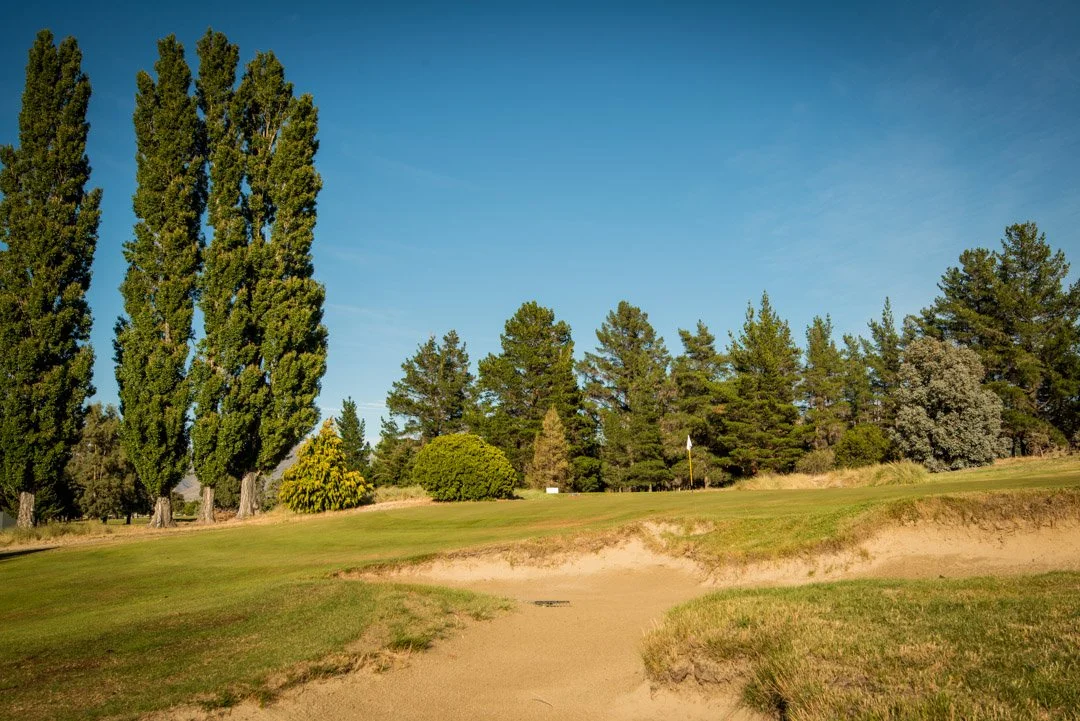 Golf course with sand bunker, green grass, and tall trees under a clear blue sky.
