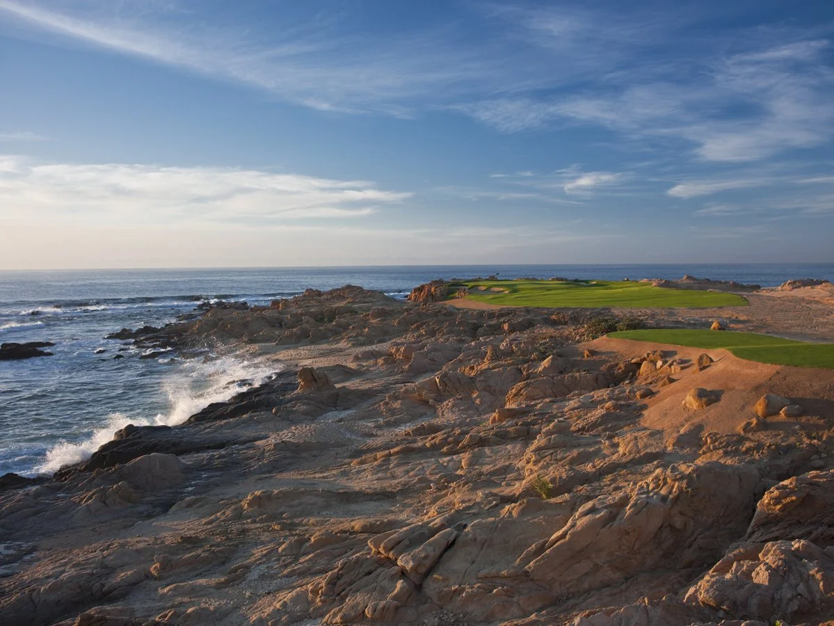 Rocky coastline with waves crashing and a golf course in the distance under a blue sky with scattered clouds.