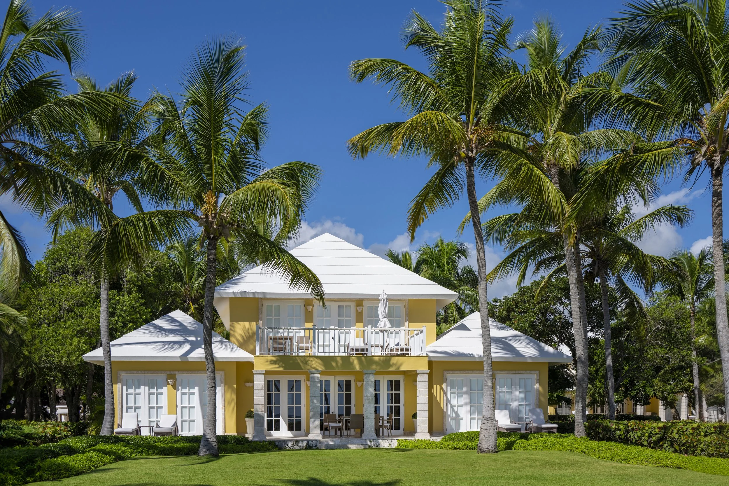 Yellow house with white roof surrounded by tall palm trees and lush green grass on a sunny day.