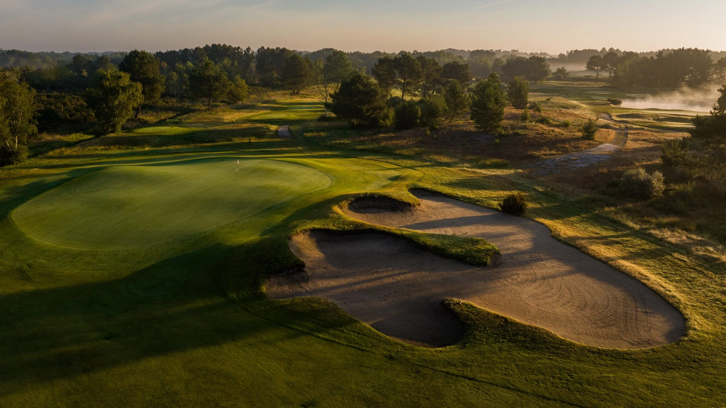 Aerial view of a golf course at sunrise with greens, sand bunkers, and surrounding trees.