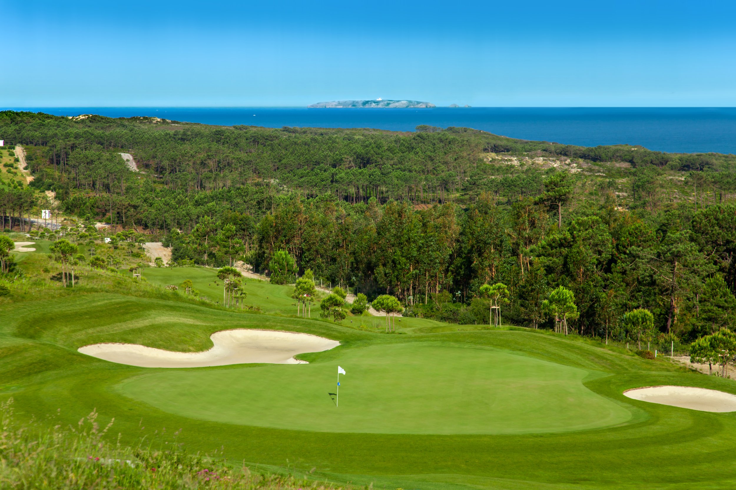 A golf course with sand bunkers and a flagstick on the green, surrounded by lush trees, overlooking the ocean and a distant island under a clear blue sky.